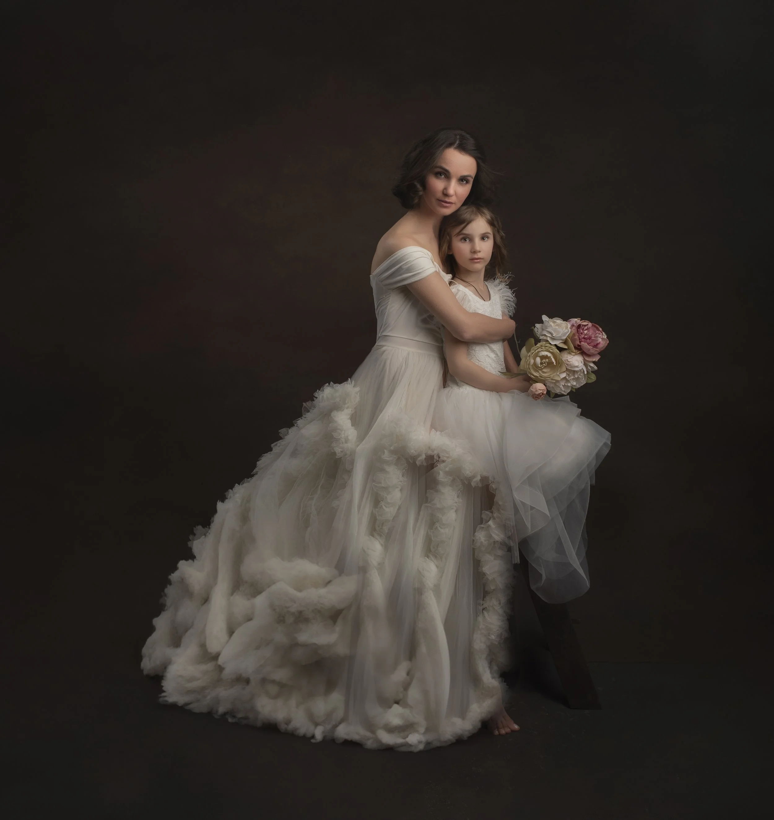 A woman and a young girl in wedding dresses posing together, with the woman holding a bouquet of flowers, against a dark studio background.