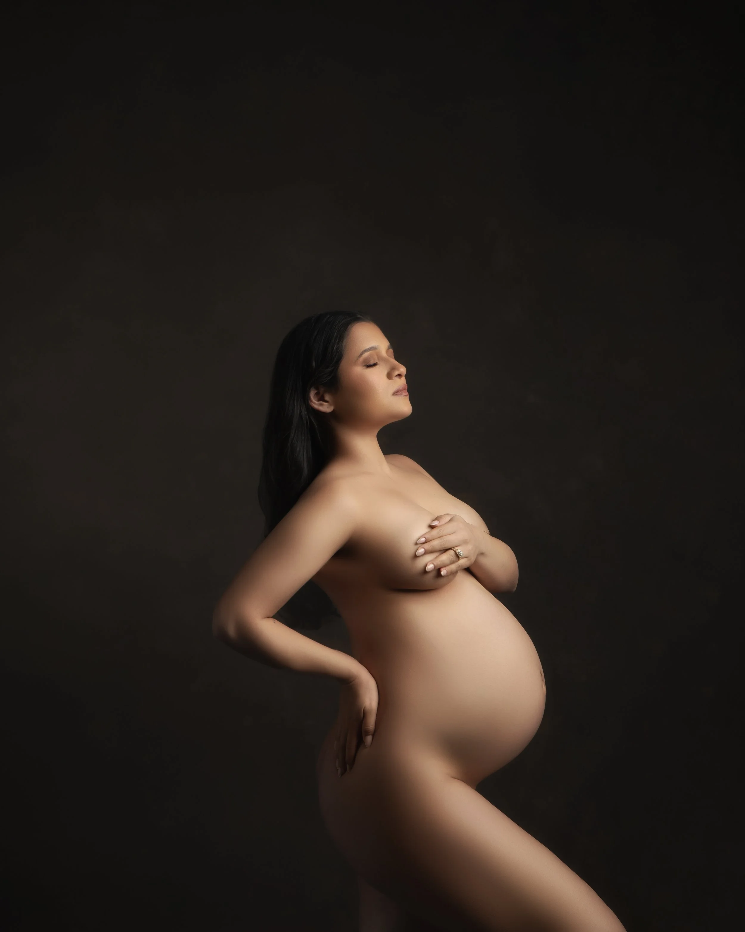 Nude pregnant woman with long dark hair posing against a dark background.