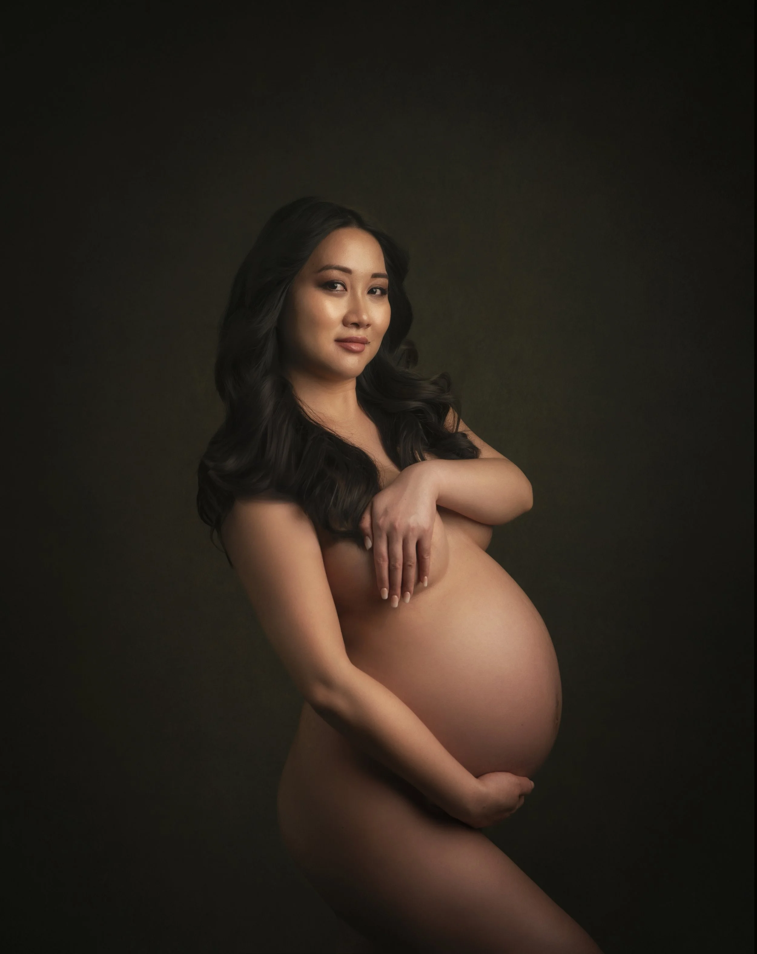 A pregnant woman with long dark hair poses topless against a dark background, cradling her baby bump with one hand and resting the other on her shoulder.