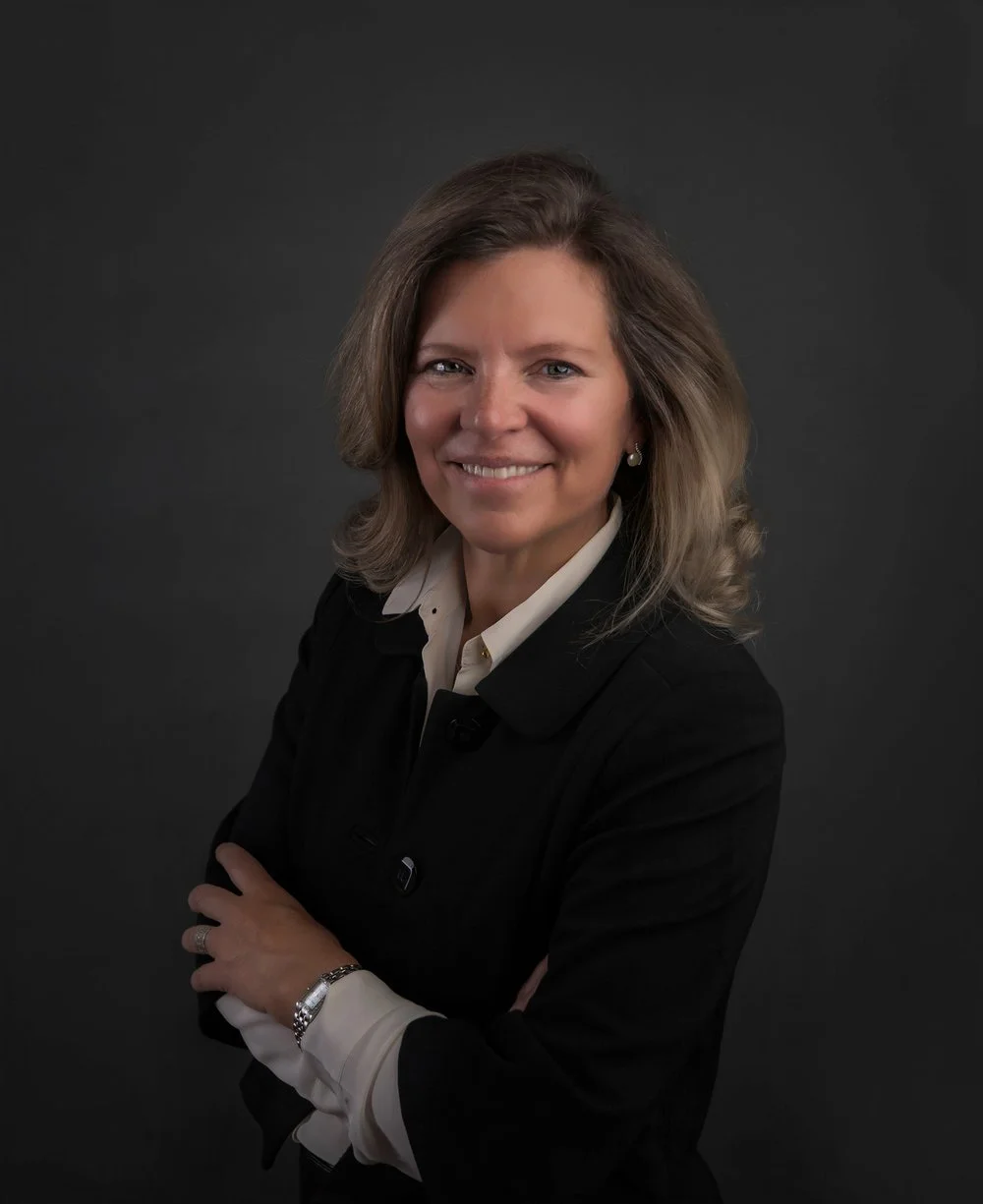 Professional headshot of a woman with shoulder-length light brown hair, wearing a black blazer over a white shirt, smiling against a dark gray background.