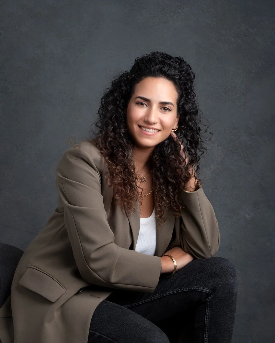 Headshots by Ally, A young woman with curly dark hair smiling and sitting against a dark, textured background. She is wearing a taupe blazer over a white top, with jewelry including earrings and a necklace.