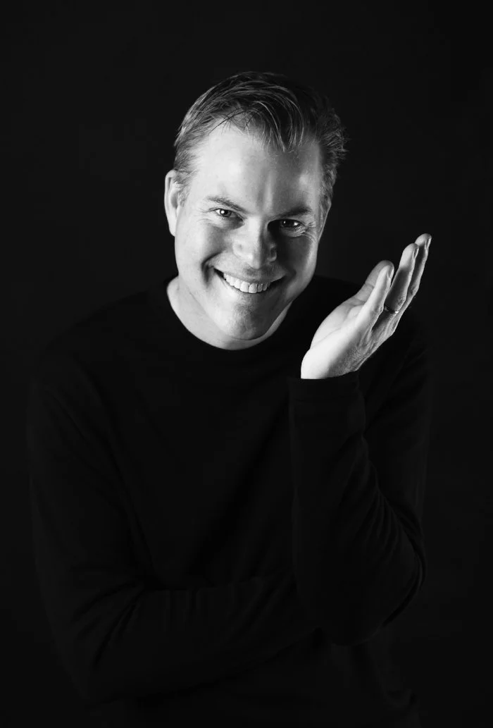 Black-and-white portrait of smiling man with slicked-back hair, wearing a dark long-sleeve shirt, posing with his hand to his face against a dark background.