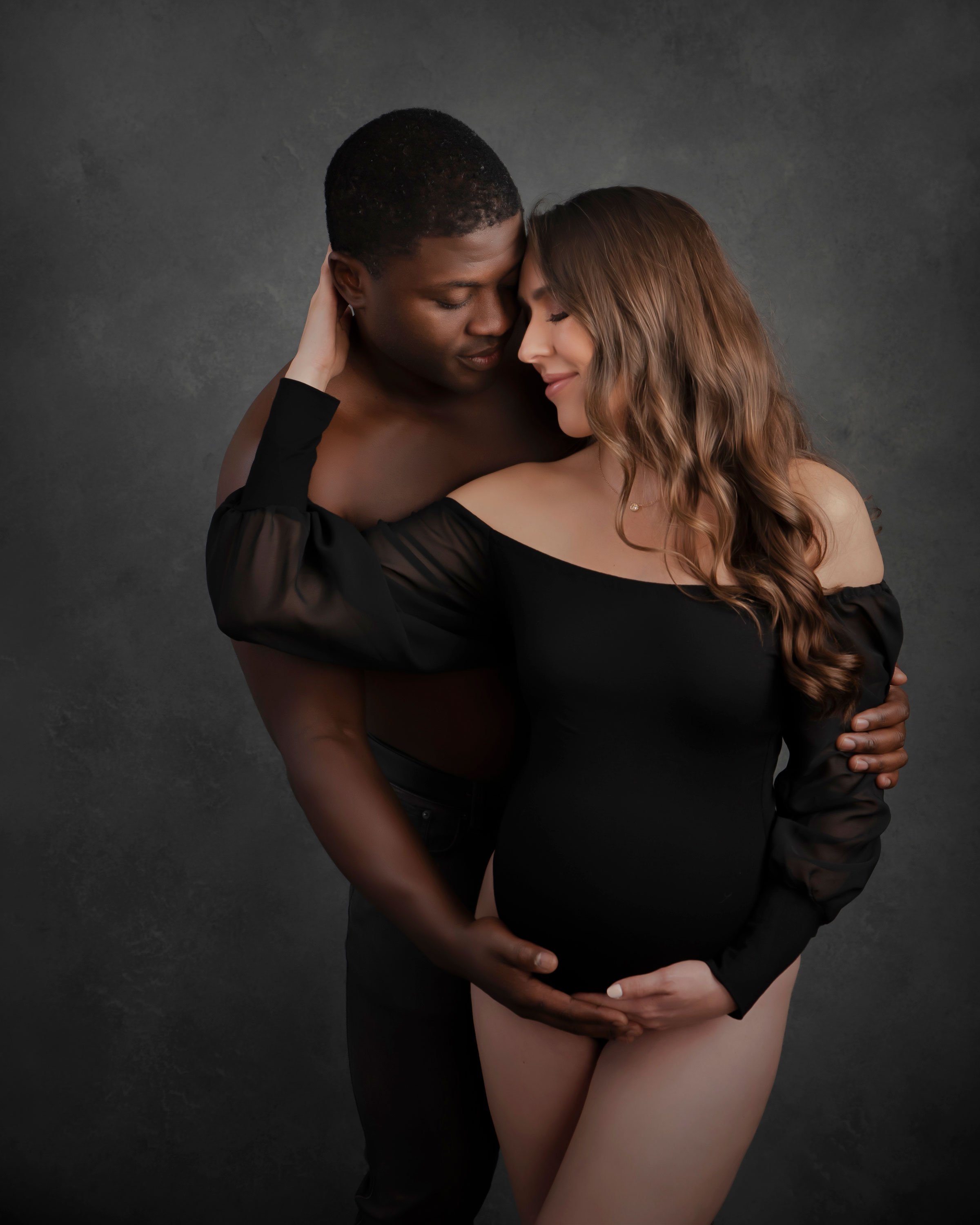 A couple embraces, with the woman pregnant, both eyes closed, in a dark background.