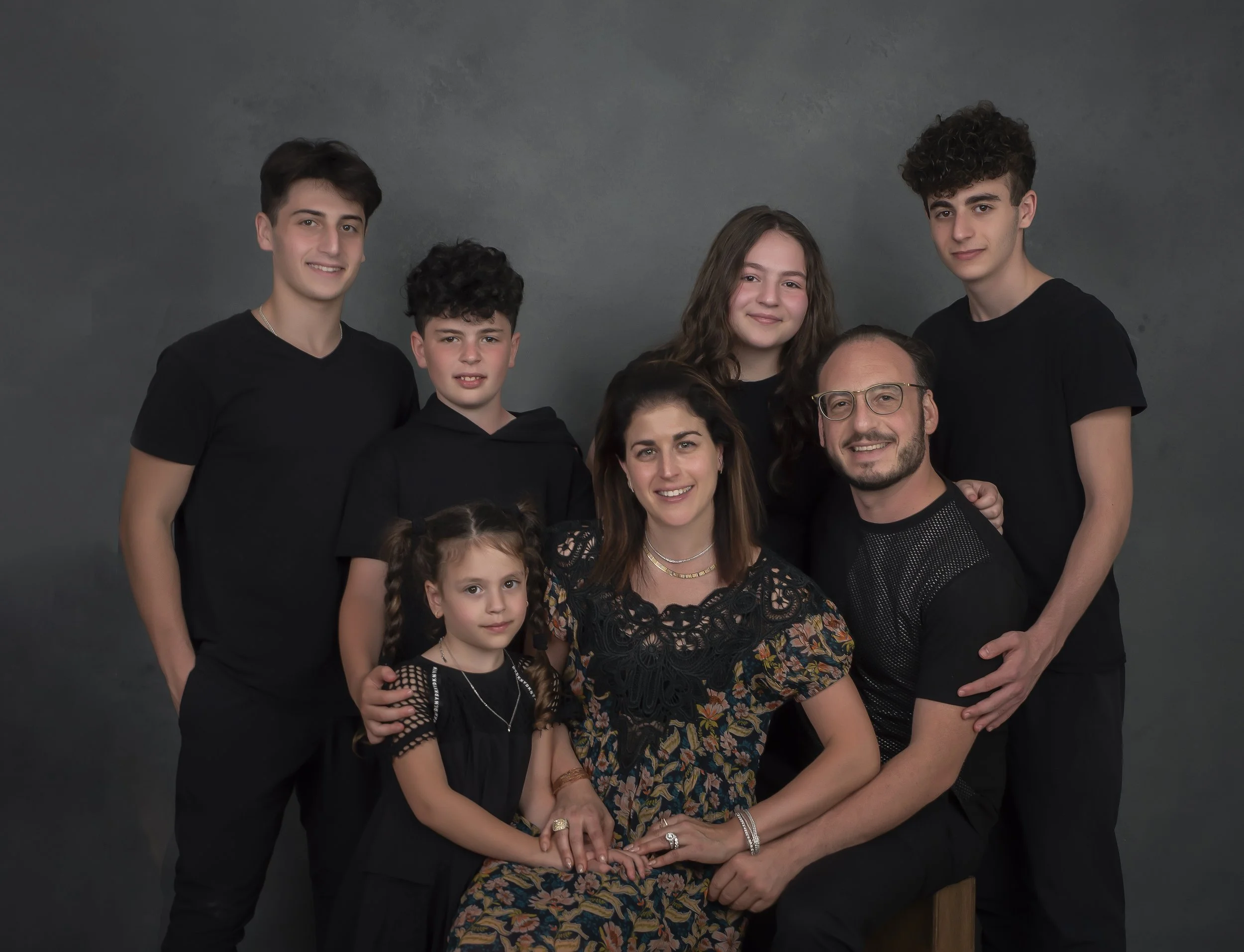 A family portrait of eight people, including a man, a woman, and six children, all dressed in black clothing, posing against a dark gray background.