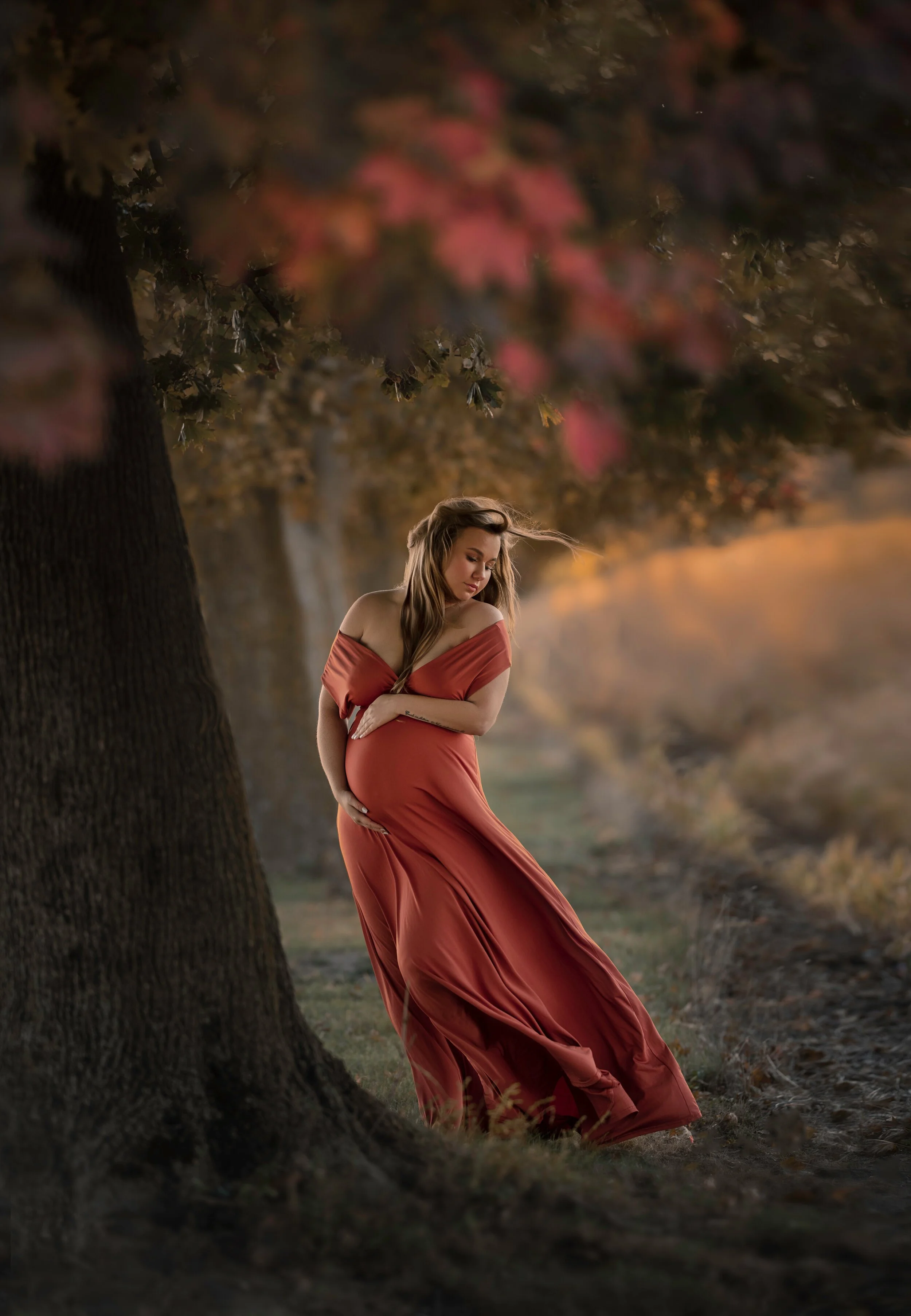 A pregnant woman in a flowing red-orange dress standing outdoors next to a large tree during sunset, with autumn trees and a soft blurred background.