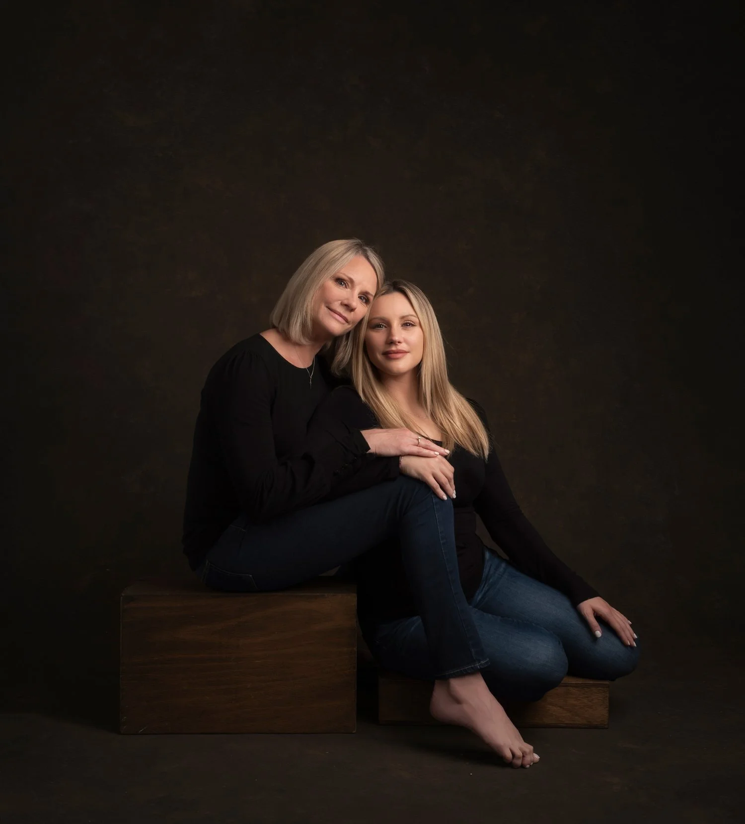 A mother and daughter sitting together, both wearing black tops and blue jeans, against a dark studio backdrop. The mother has blonde hair and the daughter has long blonde hair, sharing a warm moment with the daughter sitting on the mother's lap and 