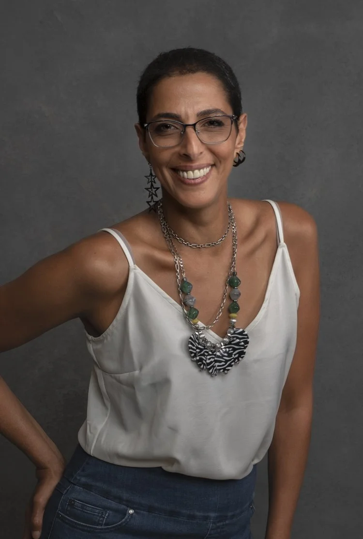 A woman with short dark hair, glasses, and star-shaped earrings, smiling at the camera, wearing a white tank top and layered necklaces against a gray background.