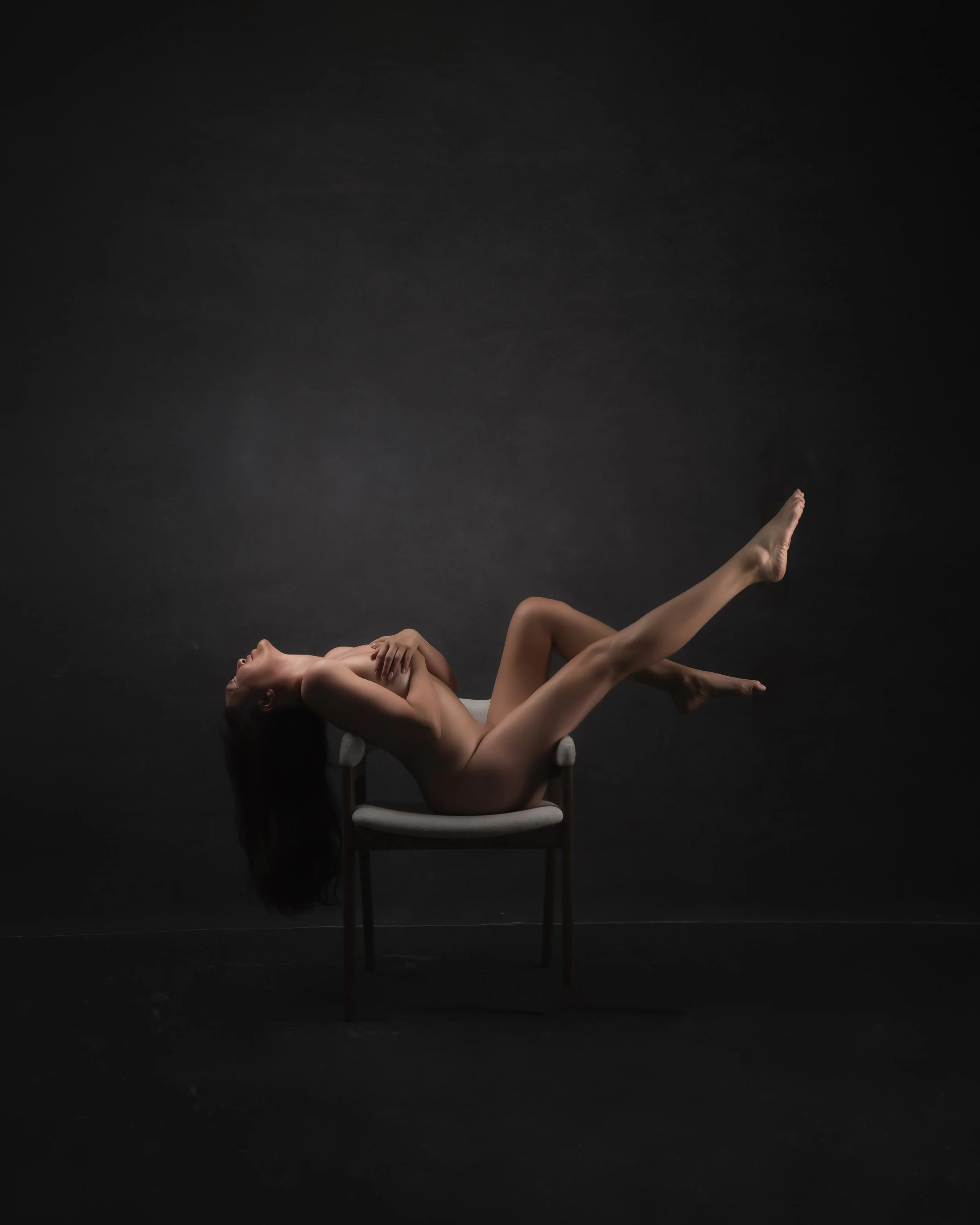 A nude woman with long dark hair reclining on a chair against a dark background, with her head tilted back and one leg extended upward.