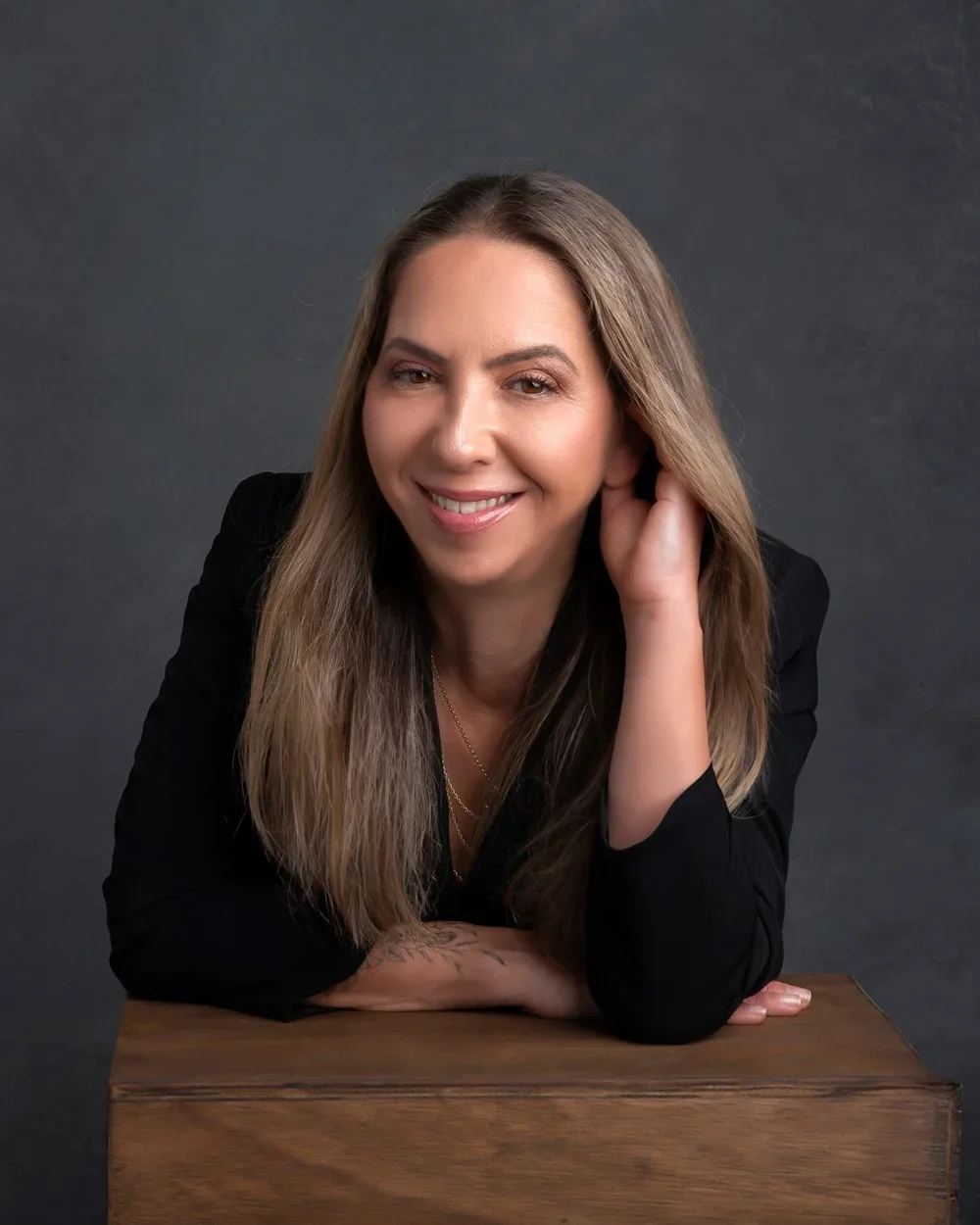 Photoshoot by AllyA smiling woman with long, light brown hair, wearing a black blazer, resting her head on her hand, leaning on a wooden block against a dark gray background.
