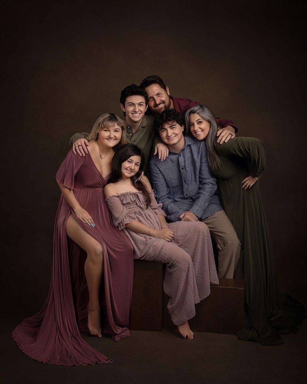 Group of seven diverse people, including young adults and older adults, posing closely together with smiles against a dark brown background, some sitting and some standing.
