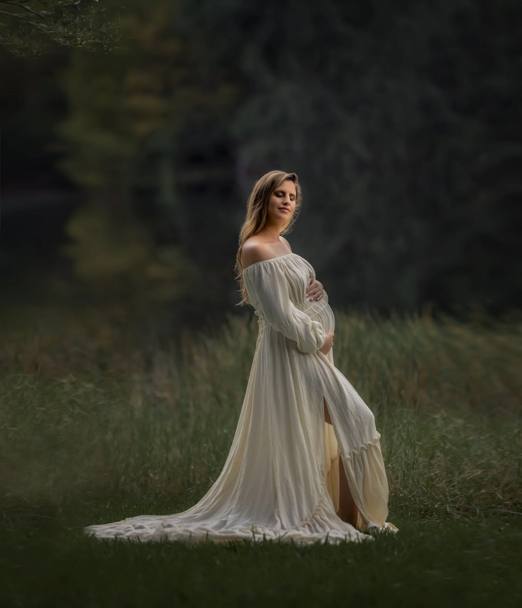 Pregnant woman in a flowing cream-colored dress standing on grass near a dark wooded area, gently cradling her baby bump with a serene expression.