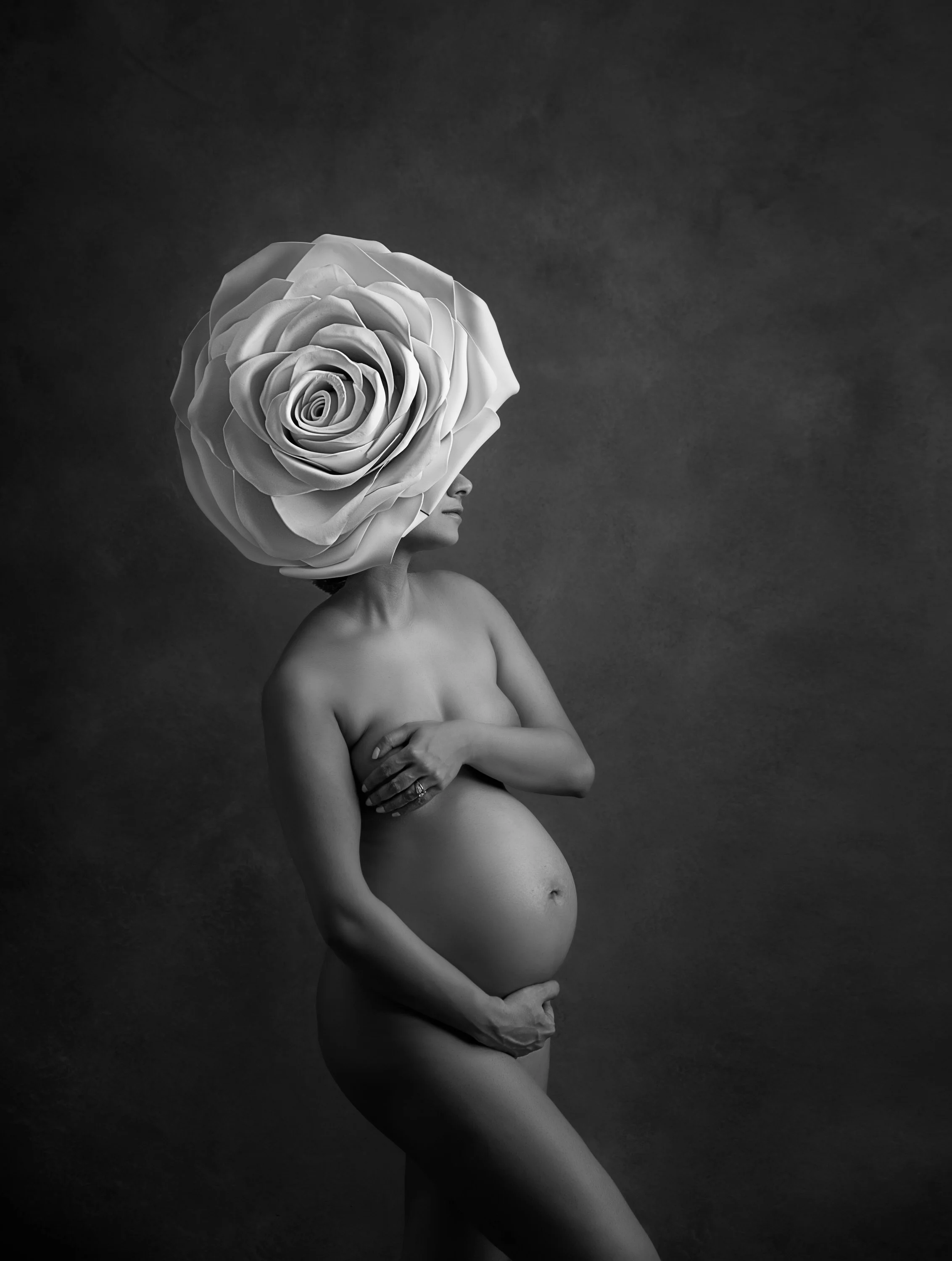 A black and white photo of a nude woman with a large, blooming rose on her head, covering her eyes. She is holding her pregnant belly with both hands, standing against a plain dark background.