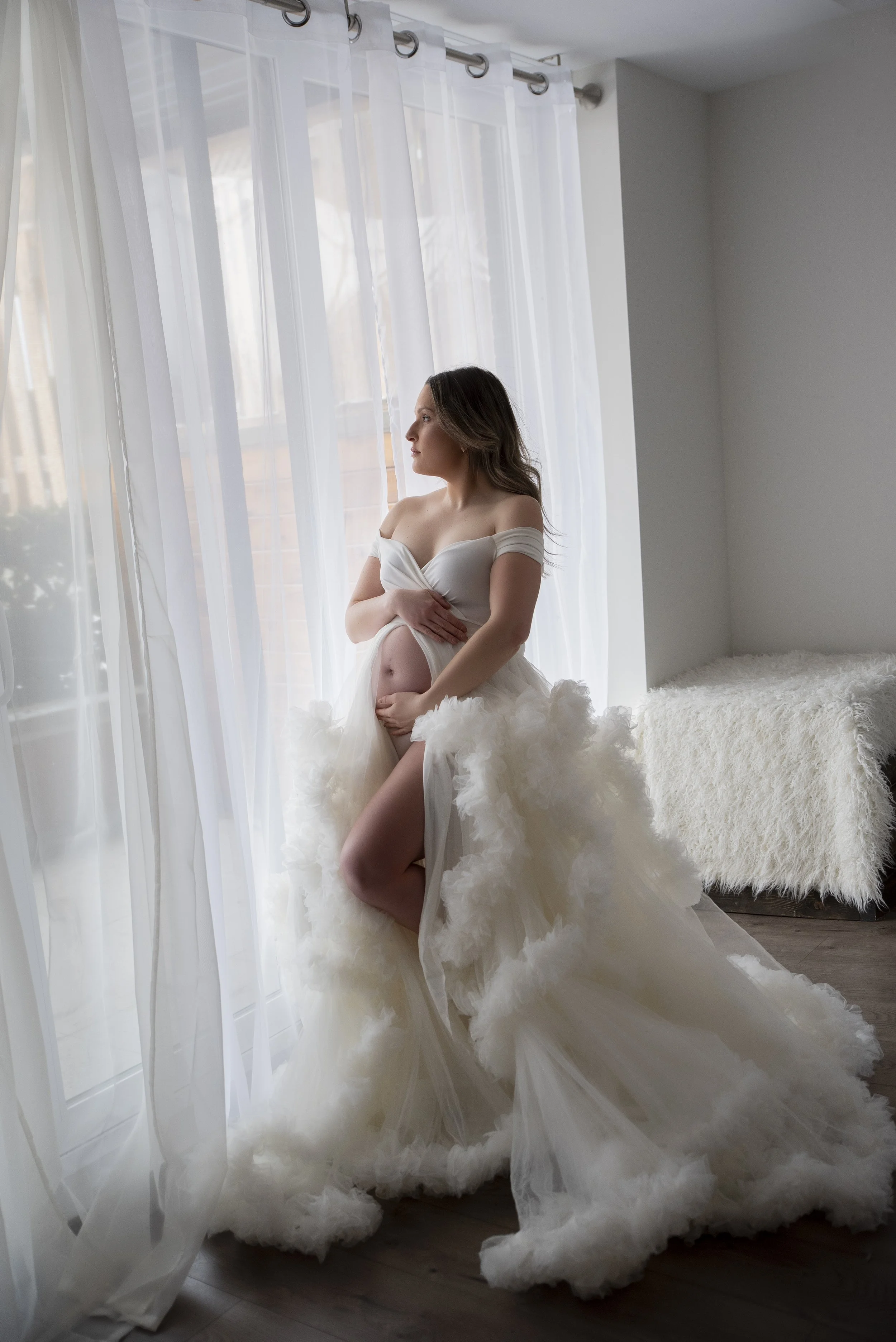 A pregnant woman in a white wedding dress standing by a window with sheer curtains, looking outside, with one hand on her belly and the other lifting her dress to show her belly.