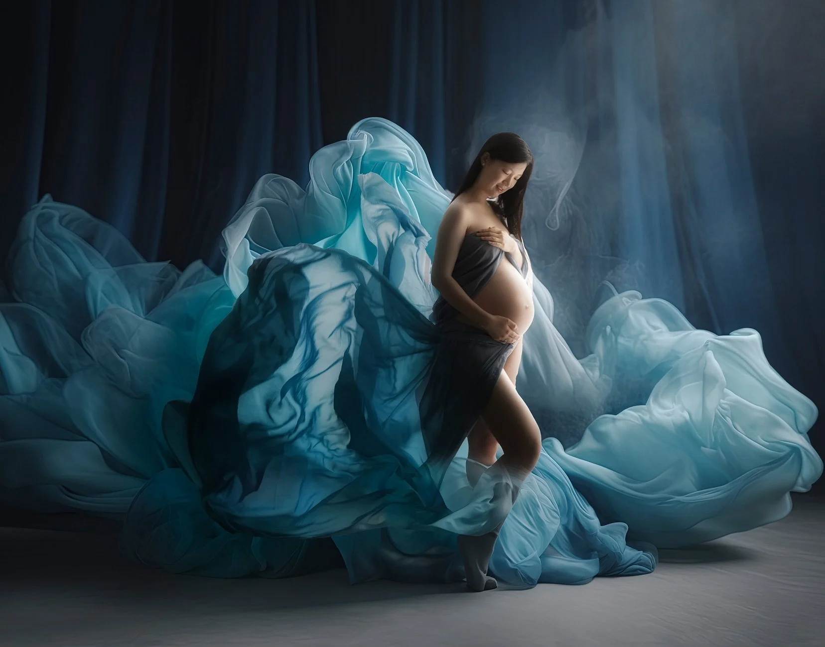Pregnant woman standing among flowing blue and white fabric with soft lighting.