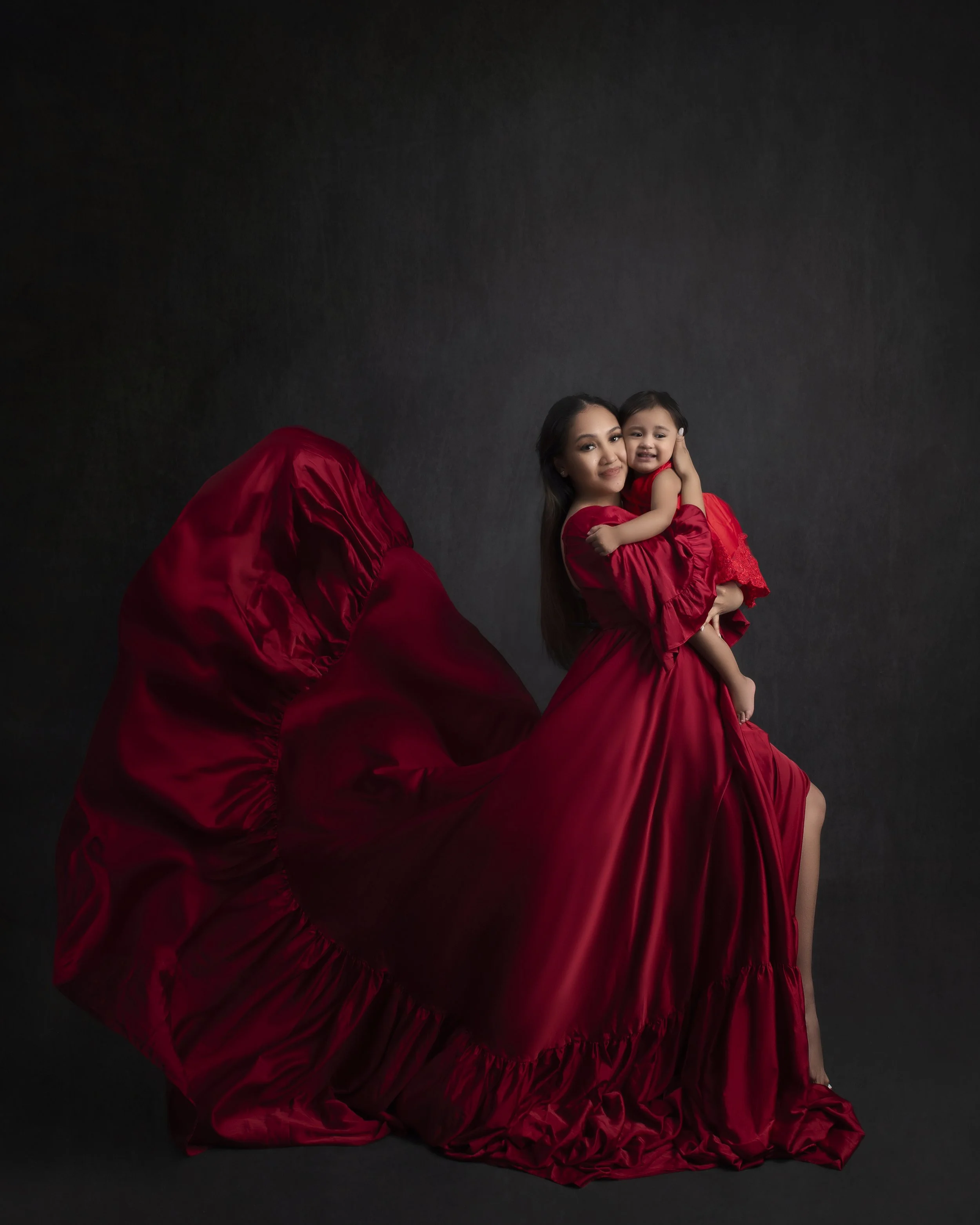 A woman holding a young girl, both dressed in red dresses, standing against a dark background.