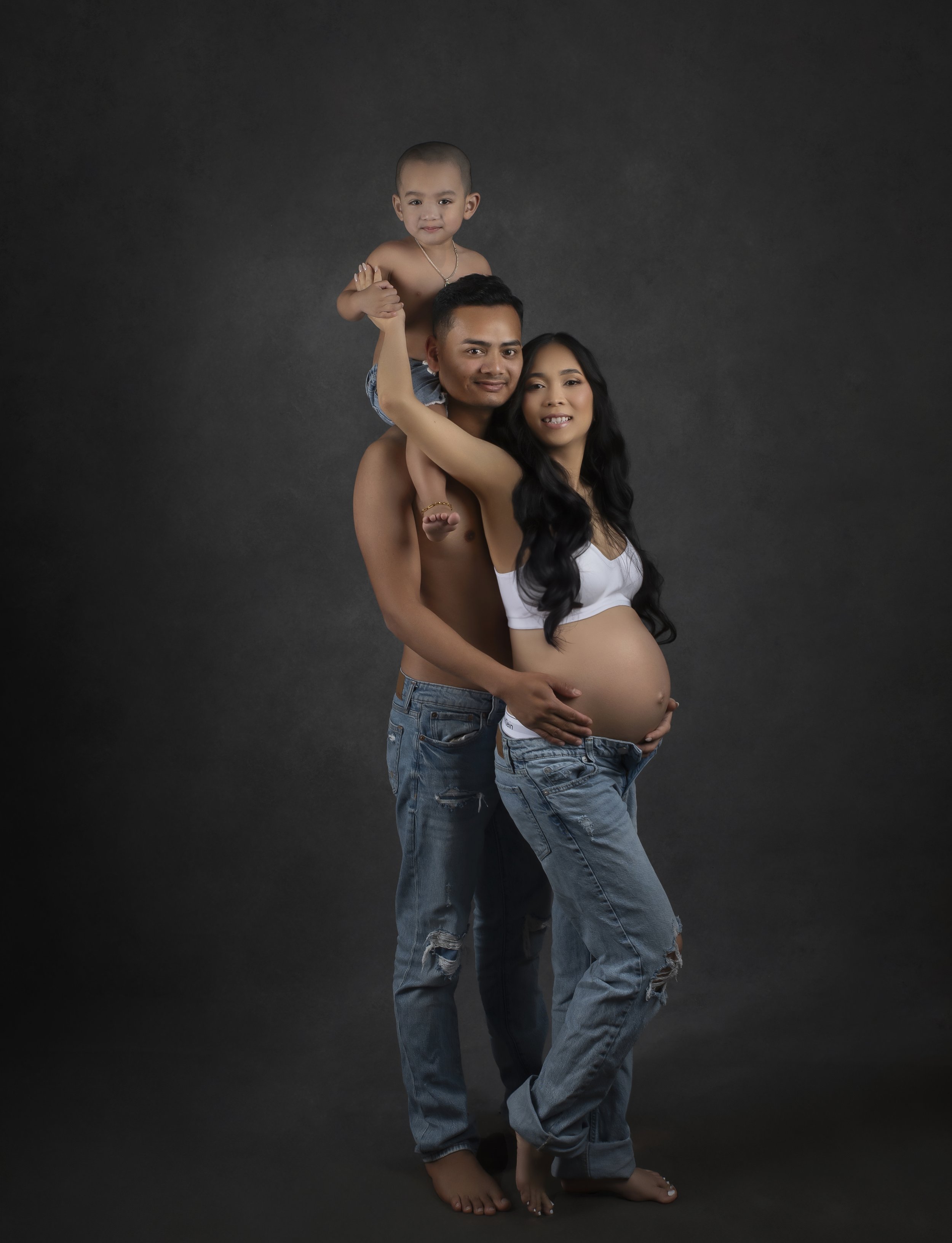 A pregnant woman, a shirtless man, and a young child pose together against a dark backdrop. The woman shows her pregnant belly, the man holds her, and the child is sitting on the man's shoulders.