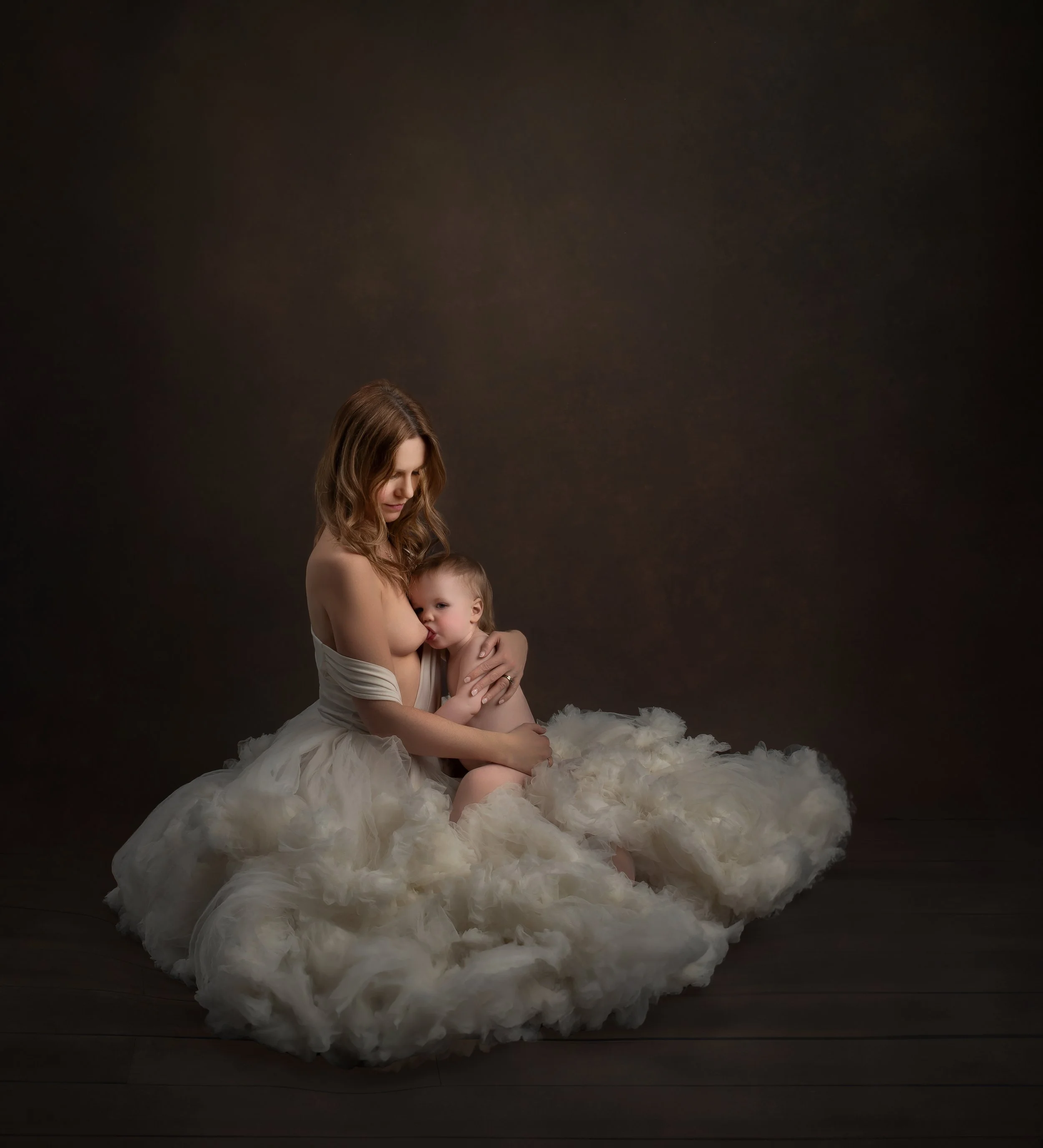 A woman with long brown hair dressed in a white off-shoulder gown sitting on a fluffy white covering, holding a young child with light brown hair, both appearing in an intimate, tender moment against a dark, neutral background.
