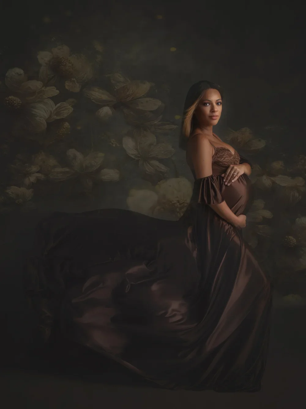 A pregnant woman in a brown dress kneels in water with floating flowers, looking at the camera.