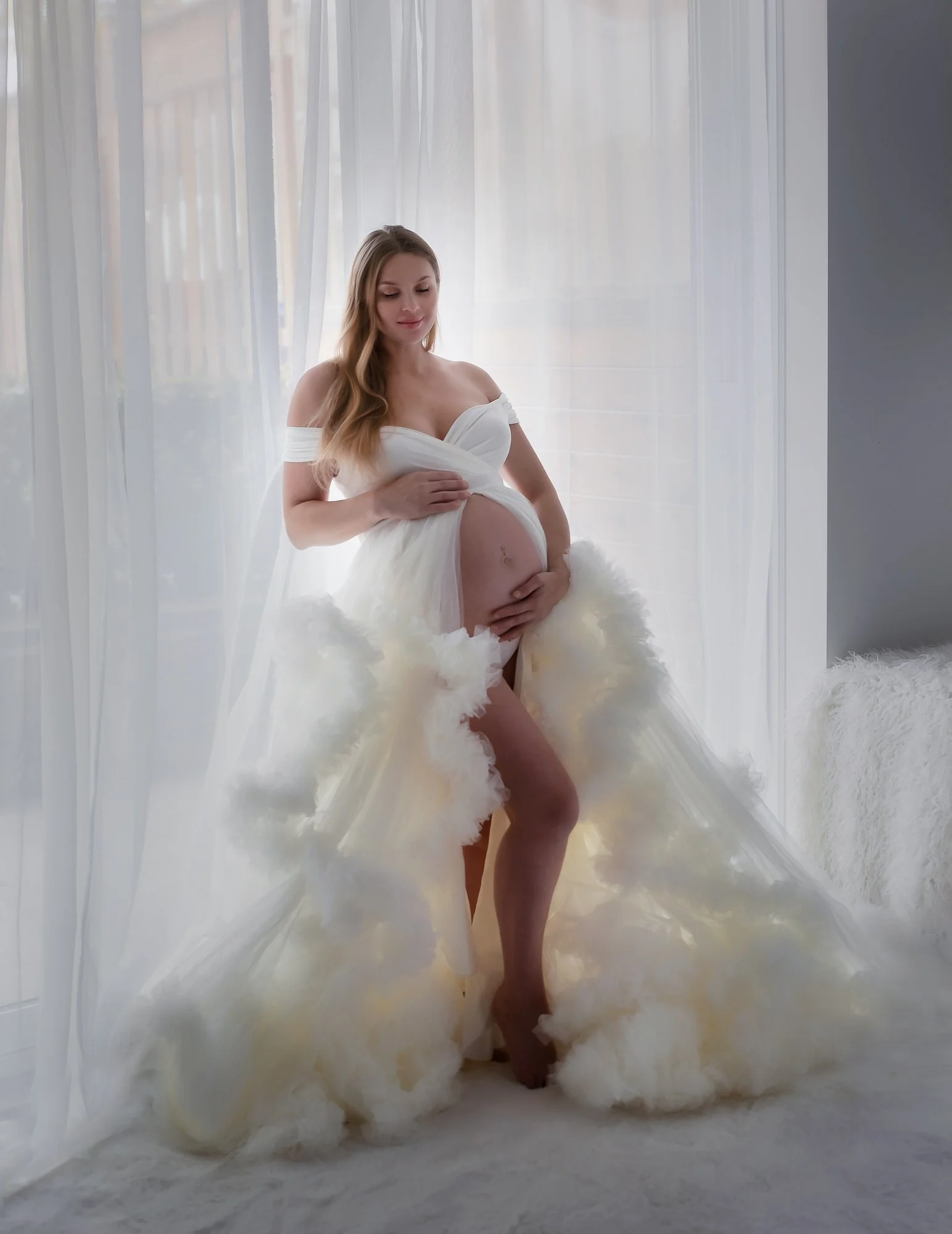 Pregnant woman in a white dress standing in front of sheer curtains, gently touching her belly with one hand and her dress with the other, on a white fluffy carpet near a window.