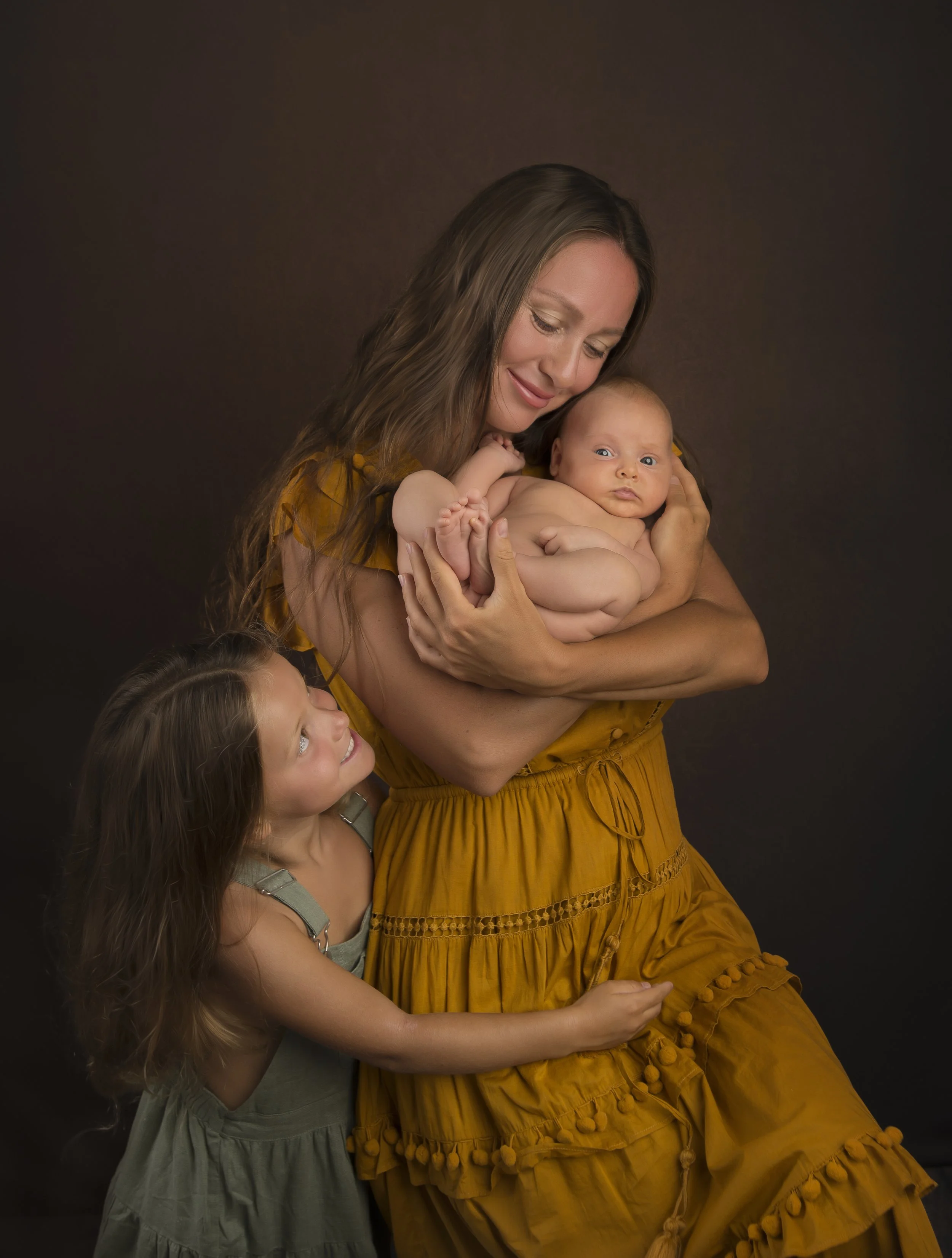A woman in a mustard yellow dress holding a newborn baby, with a young girl leaning on her and looking up at her.
