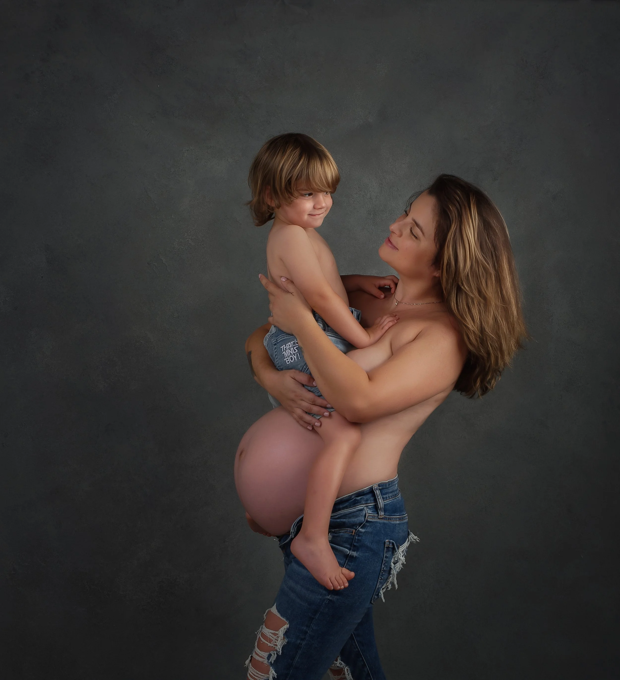 A woman with long hair, partially undressed with ripped jeans, holding a young boy with short hair who is shirtless and wearing denim shorts, against a plain dark background.
