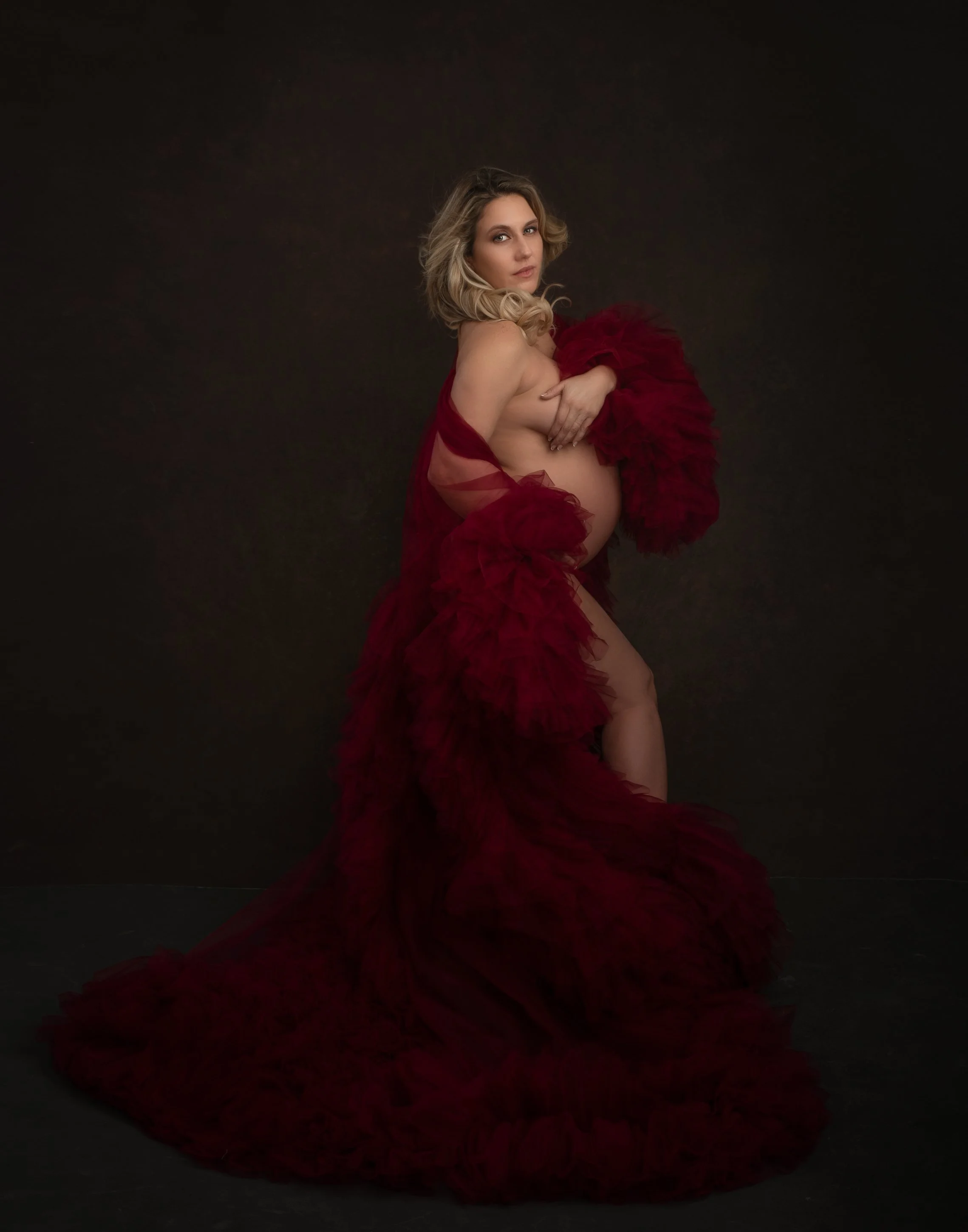 A pregnant woman draped in a flowing red tulle fabric against a dark background.