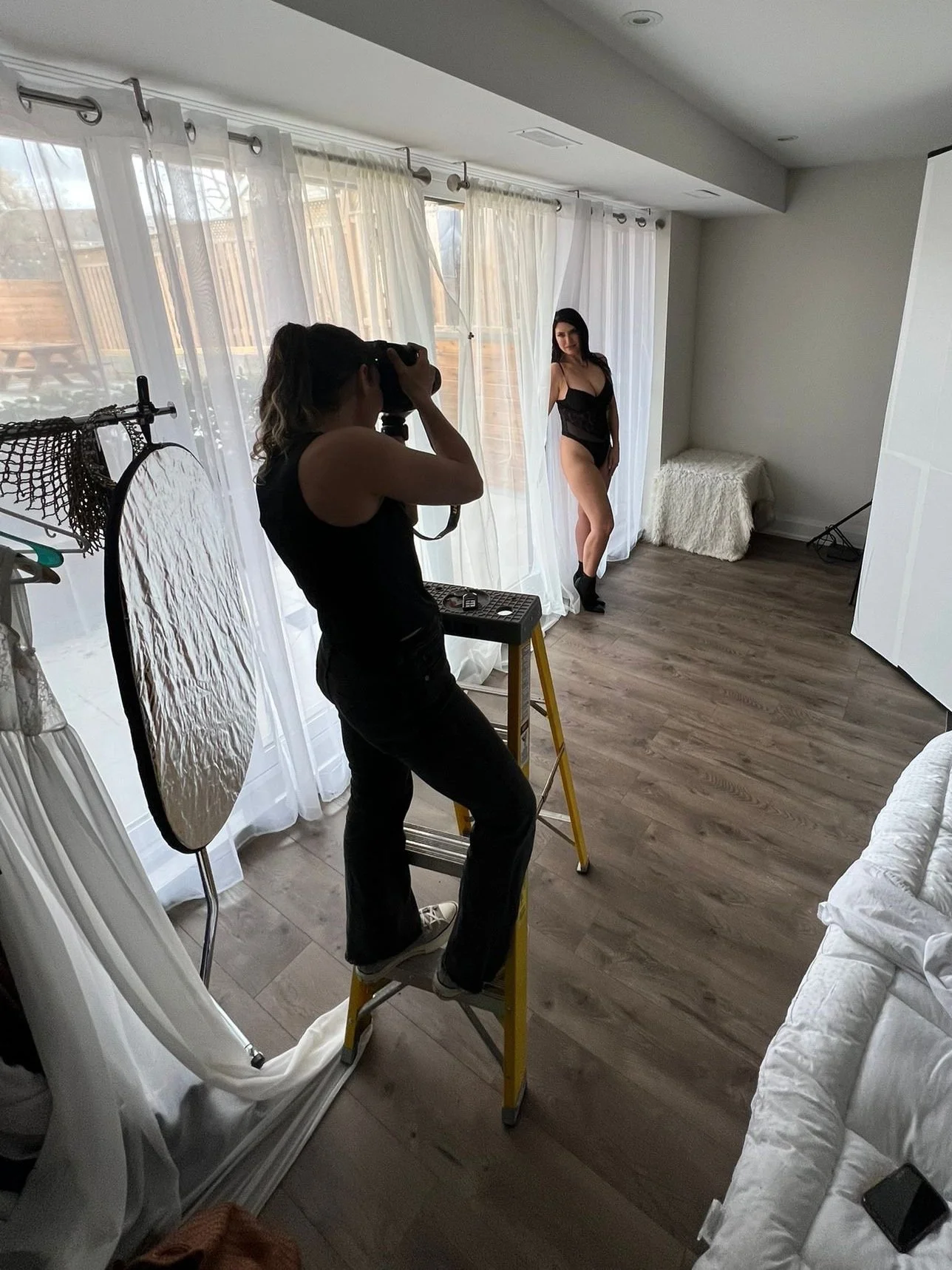 A photographer taking a photo of a woman posing in black lingerie near a window with white curtains.