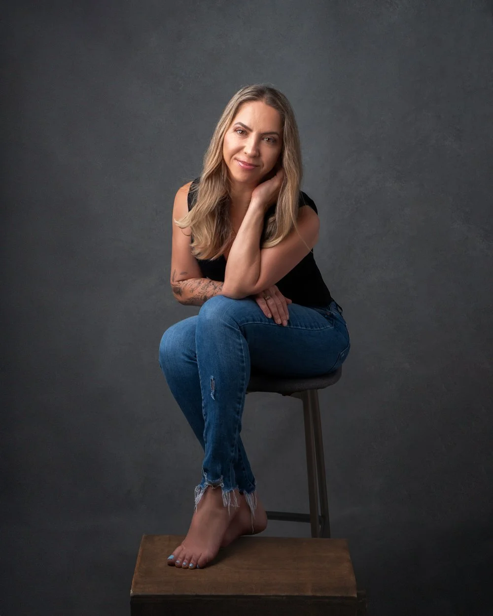 Headshots by Ally, A young woman with long blonde hair sitting on a stool against a dark gray background. She is wearing a black sleeveless top and ripped blue jeans while barefoot. She is smiling gently and resting her chin on her hand.