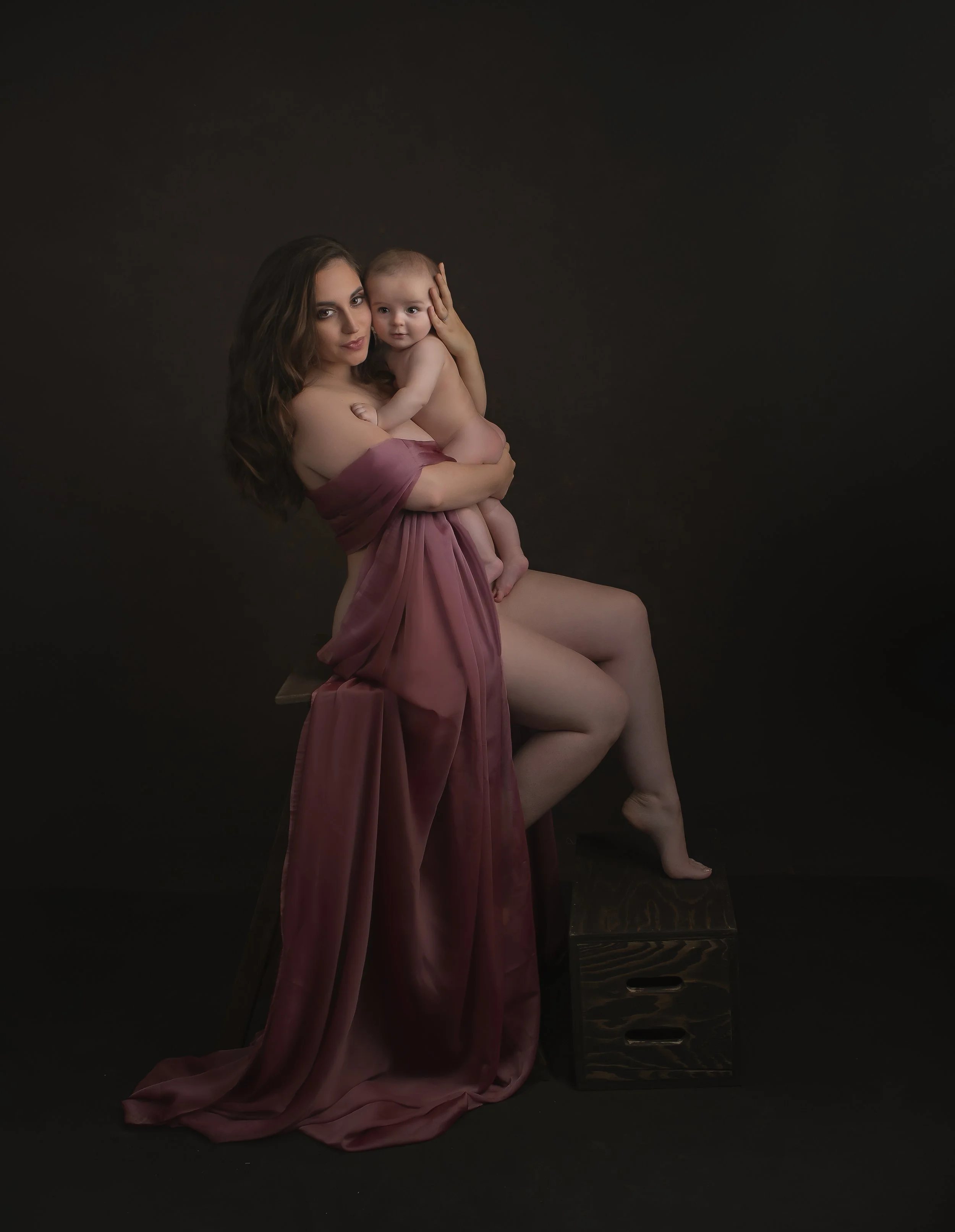 A woman holding a baby, both partially draped in pink cloth, posed against a dark background with a wooden box.
