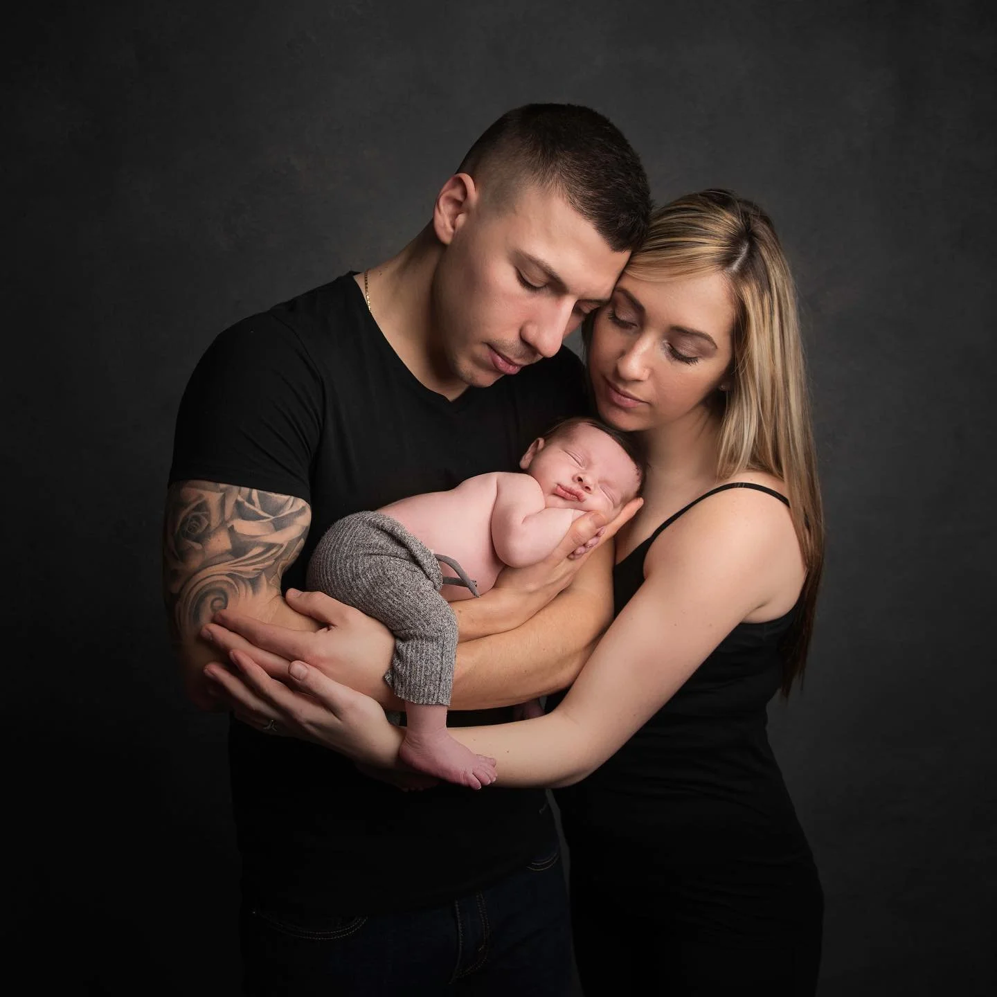 Newborn by Ally A young family of three, father, mother, and a newborn baby, posed together in front of a dark background. The father and mother share a tender moment as they hold their sleeping baby.