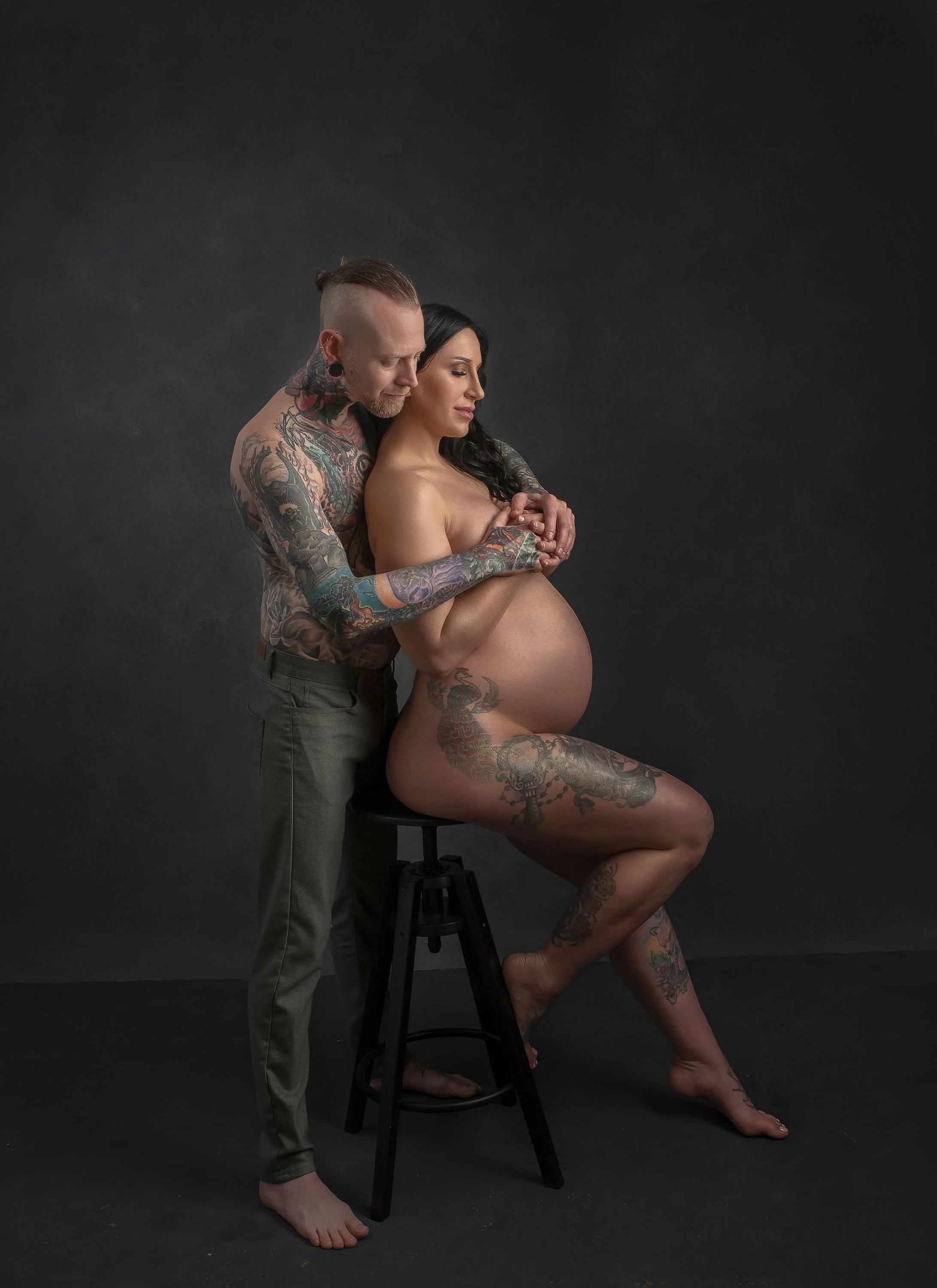 Tattooed man and pregnant woman sitting close, embracing, and touching her belly against a plain dark background.