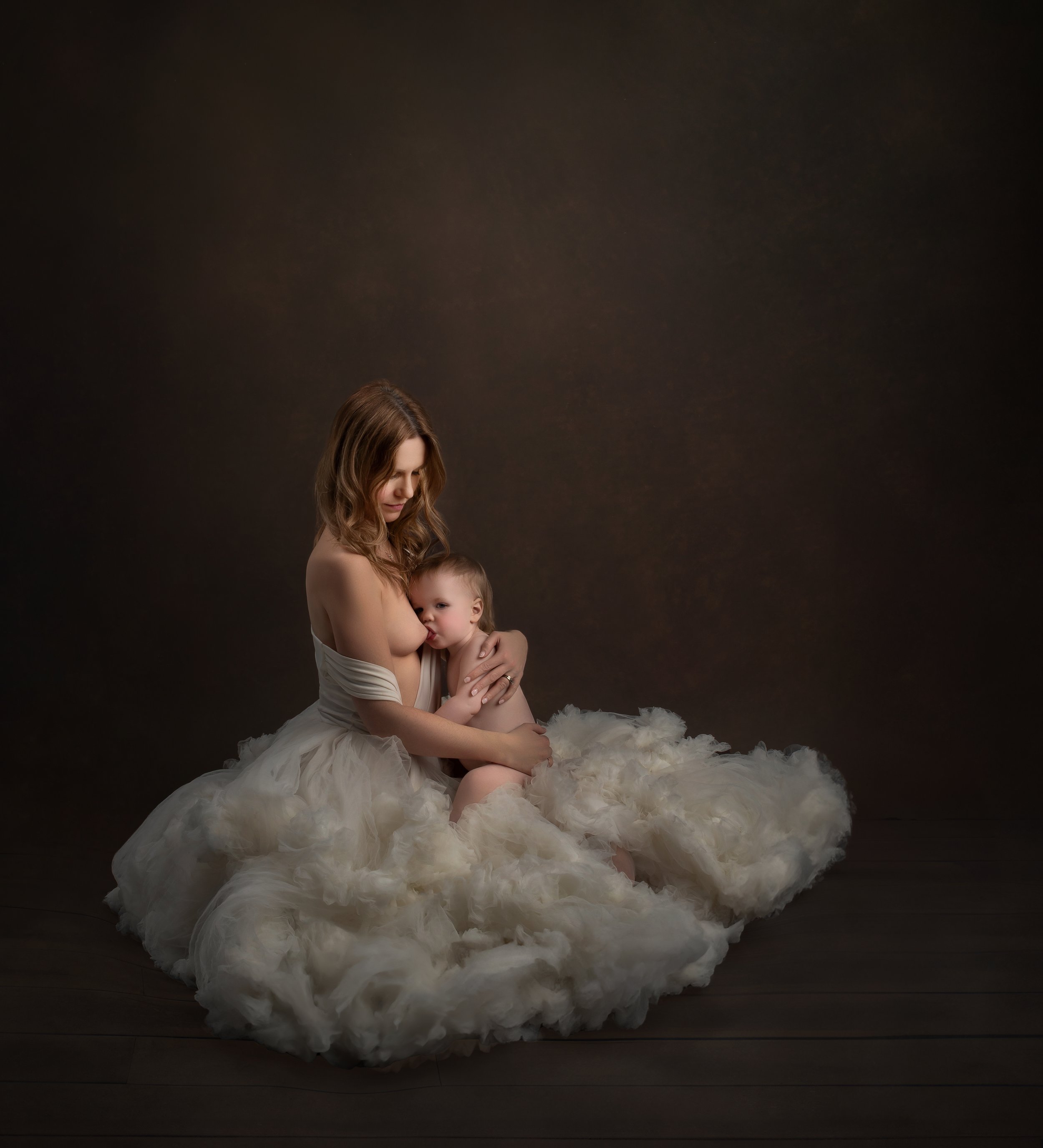 A woman and a young child sit together, with the woman holding the child and both embracing affectionately. The woman wears a flowing white dress, and the child is partially unclothed, nestled against her. The scene has a dark, neutral background, an