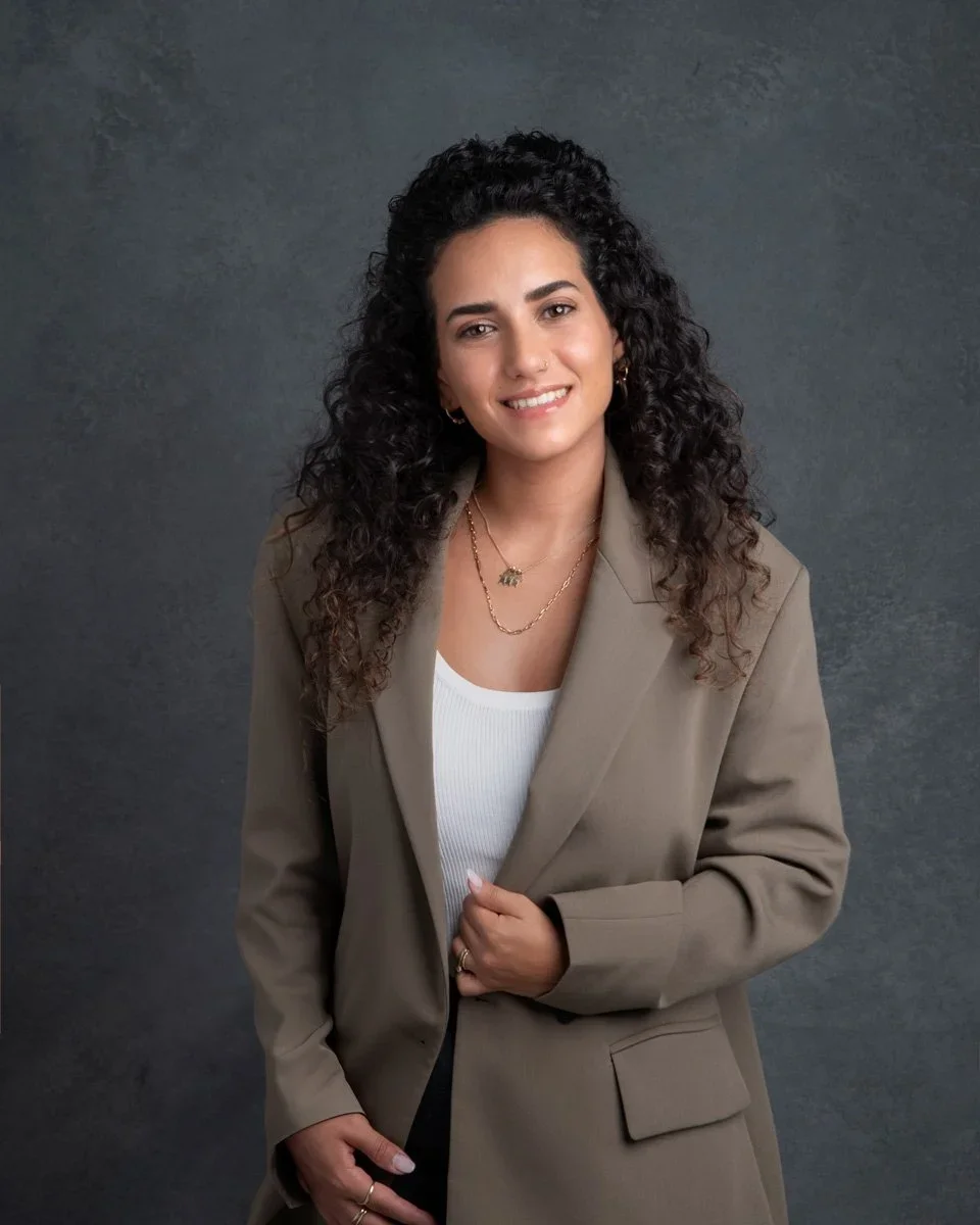 Photoshoot by Ally, Portrait of a woman with long curly dark hair wearing a beige blazer, white top, and gold jewelry, smiling at the camera against a dark gray background.