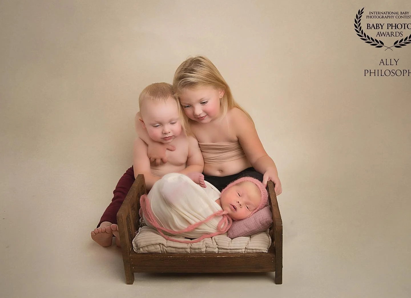 Photoshoot by AllyThree children, two older girls and a newborn baby, gather around a small wooden bed with a baby inside. The baby is wrapped in a cream blanket with pink accents, wearing a pink knit hat, and lies on a pink pillow. 