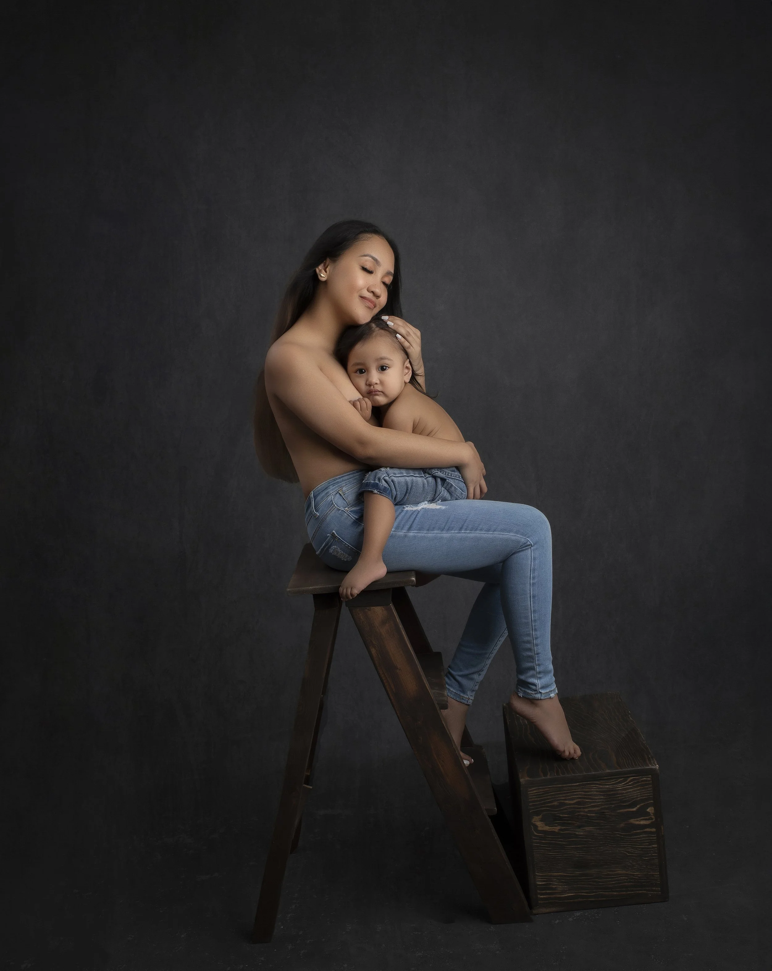 A woman holds a young girl sitting on her lap, both are shirtless, seated on a wooden stool against a dark background, creating a warm, intimate portrait.