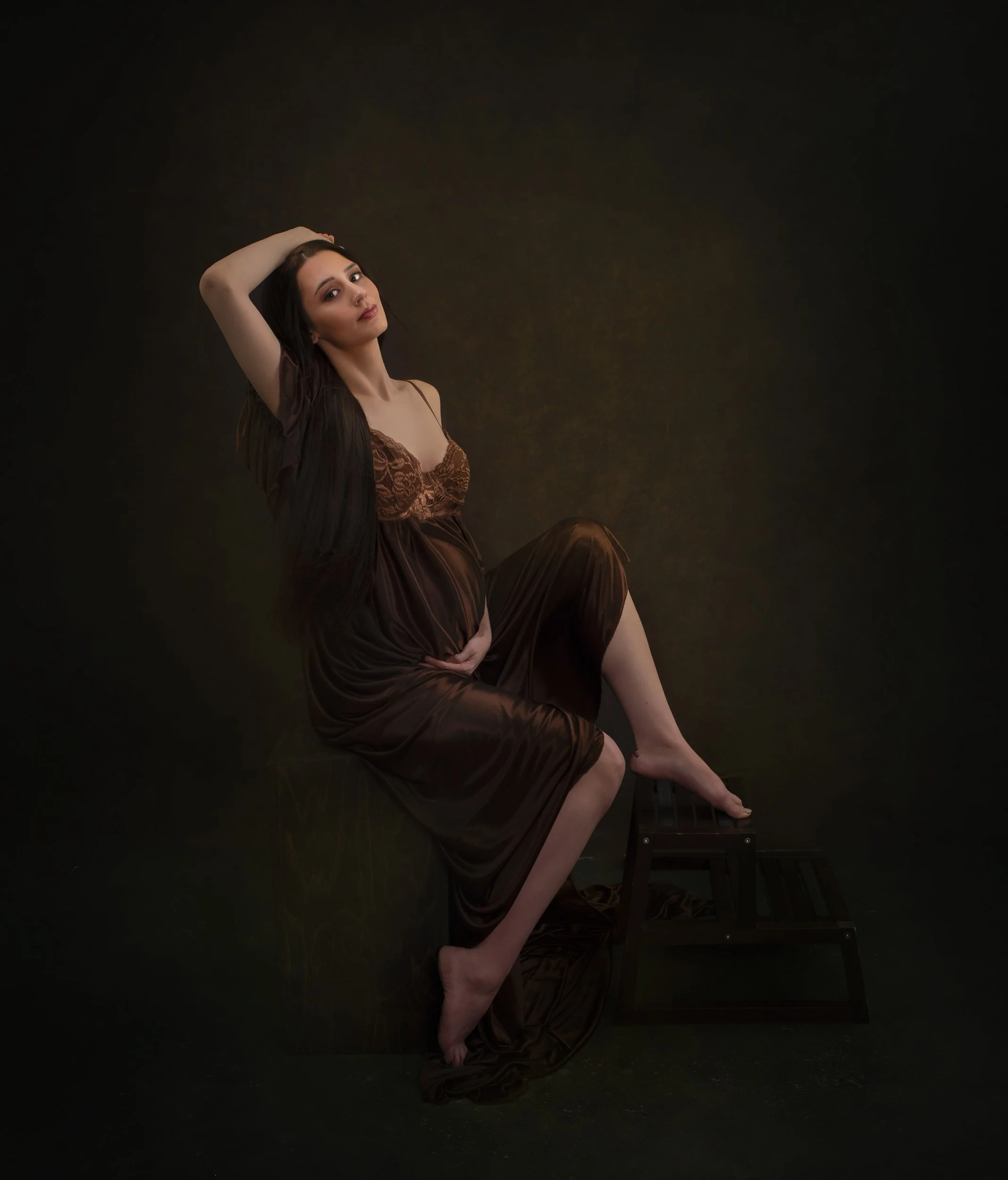 A pregnant woman in a brown satin dress sitting on a stool against a dark background, with one hand on her belly and her other arm raised above her head.