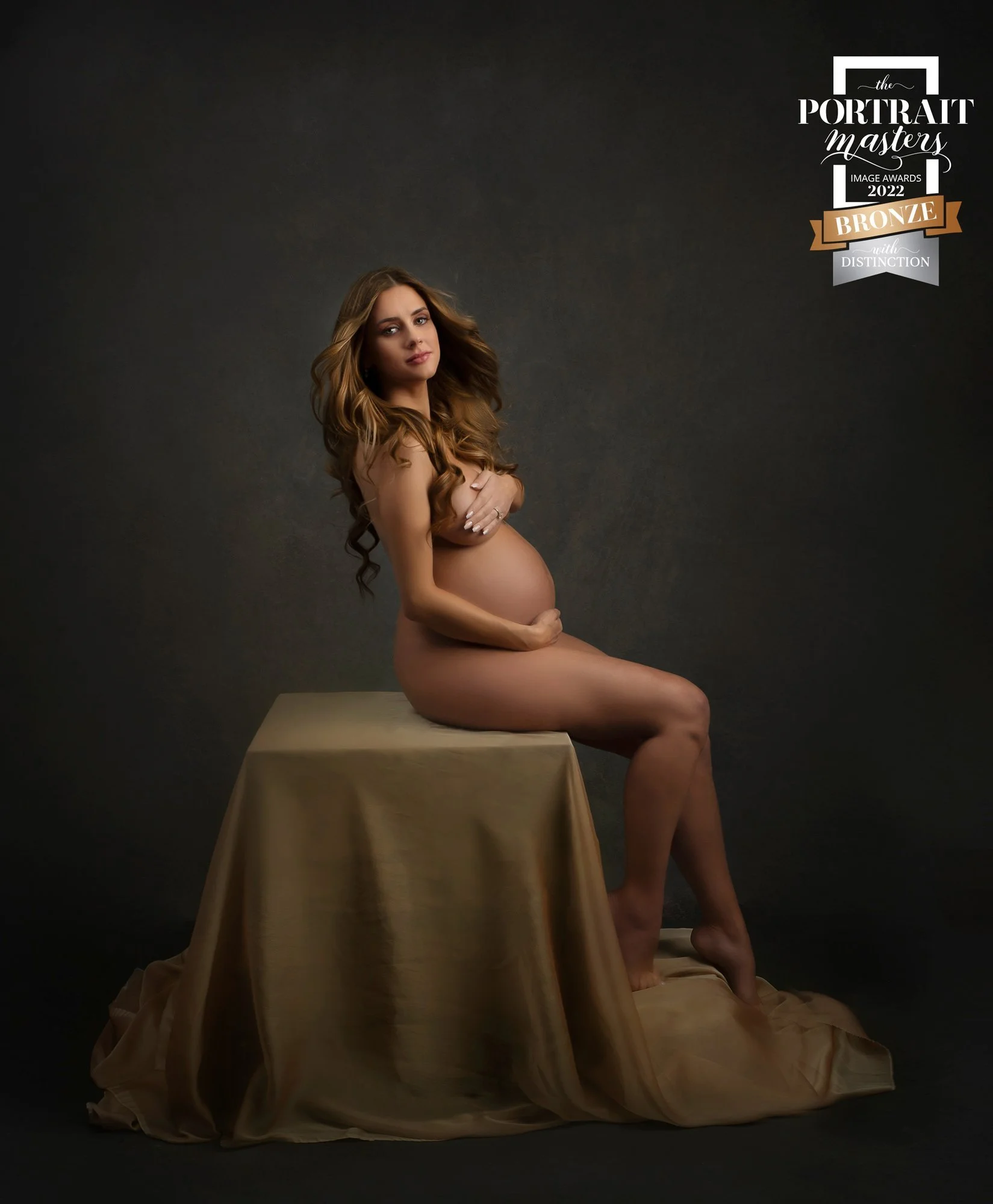 A pregnant woman with long, curled hair sitting on a gold draped stool, holding her belly with one hand and covering her chest with the other, against a dark background. There is a logo at the top right corner that reads 'The Portrait Masters 2022 Br