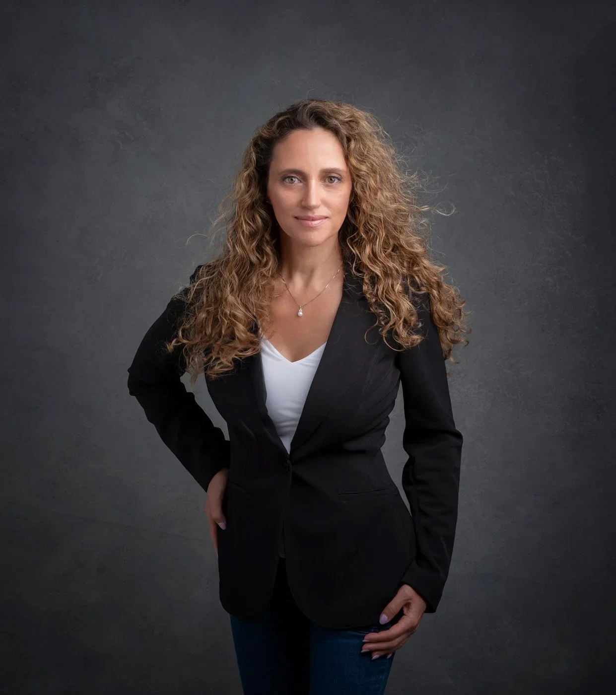 Photoshoot by Ally, A woman with curly, reddish-brown hair wearing a black blazer over a white top, standing against a dark gray background.