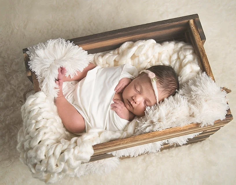 Photoshoot by Ally, A peacefully sleeping newborn baby lies on a cozy, chunky knit blanket in a wooden box, wrapped in a white swaddle and wearing a headband with a small bow.