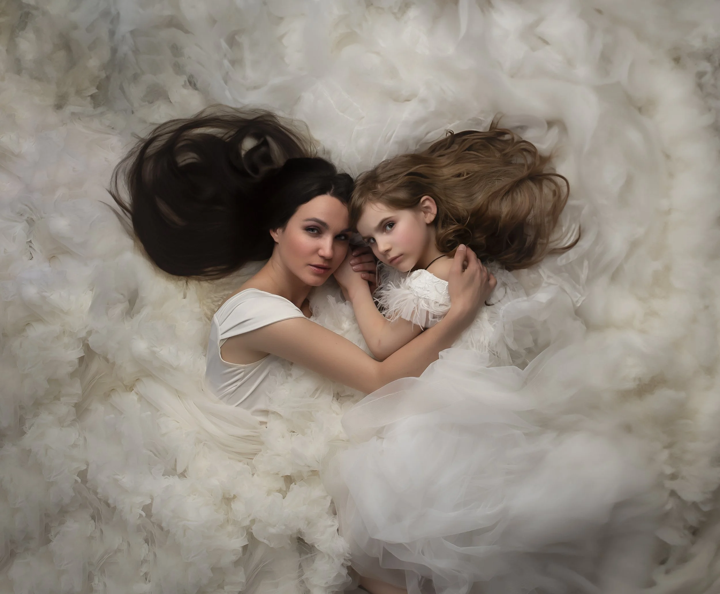 Two women, one adult and one young girl, lying down on a fluffy white surface, both dressed in white gowns, close together, looking at the camera.