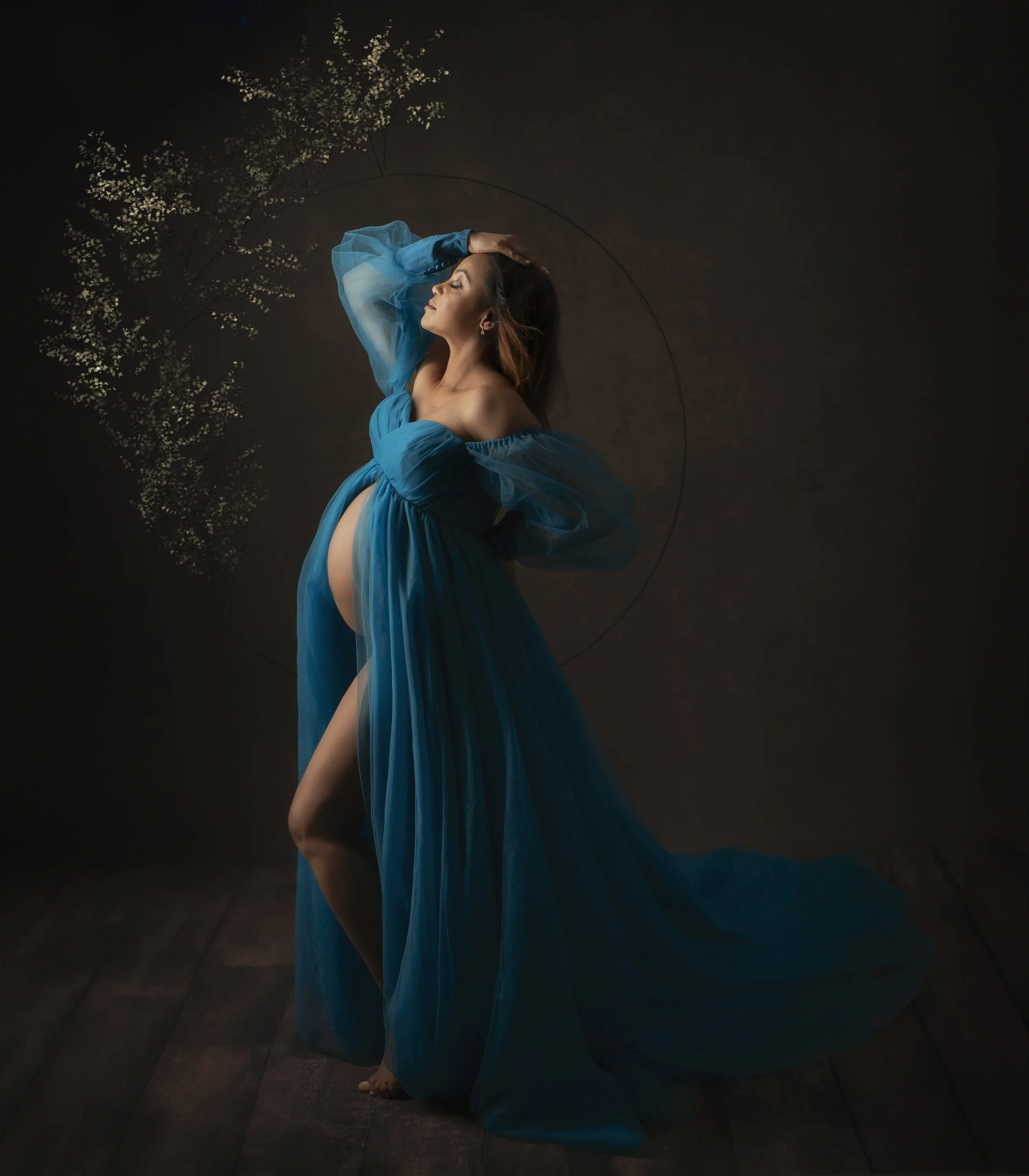 A pregnant woman in a flowing blue gown poses with one arm above her head against a dark background with floral accents.