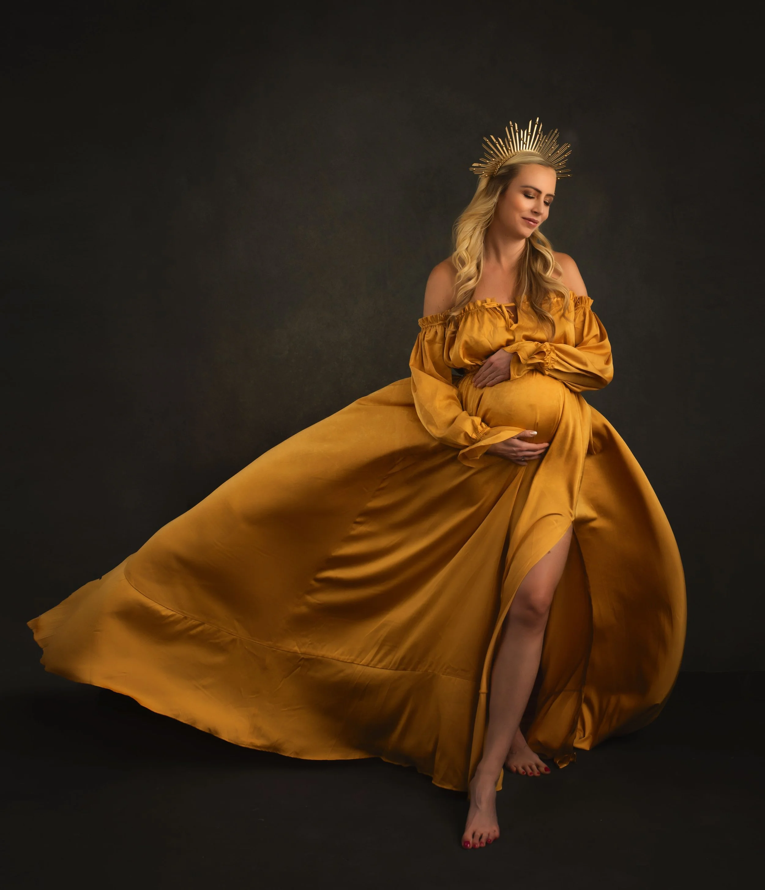 Pregnant woman in a gold gown with a matching headpiece, posed against a dark backdrop.