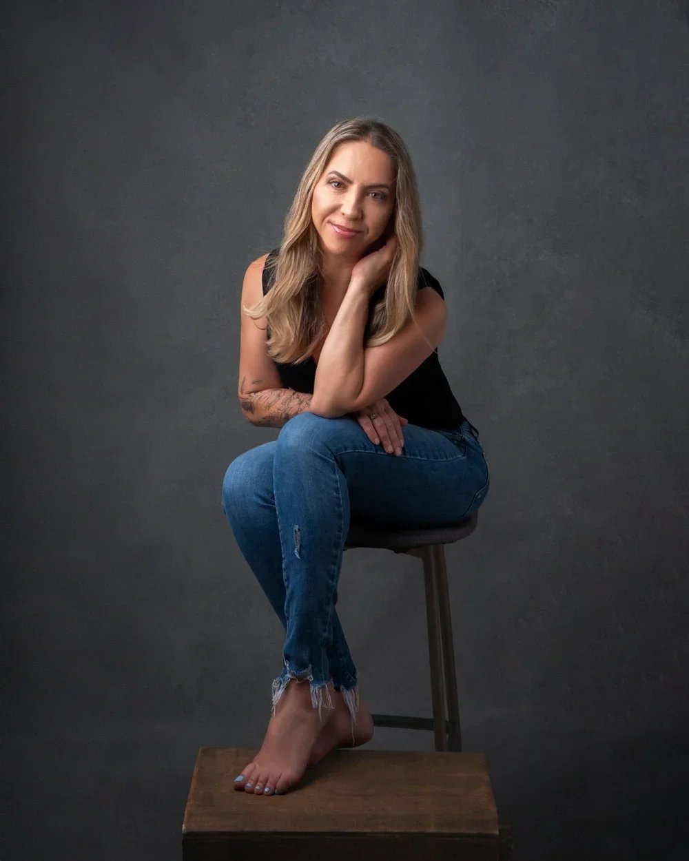 Photoshoot by AllyA woman with long blonde hair and tattoos on her arm, sitting on a stool with her legs crossed and barefoot, wearing a black sleeveless top and ripped blue jeans, against a dark textured background.