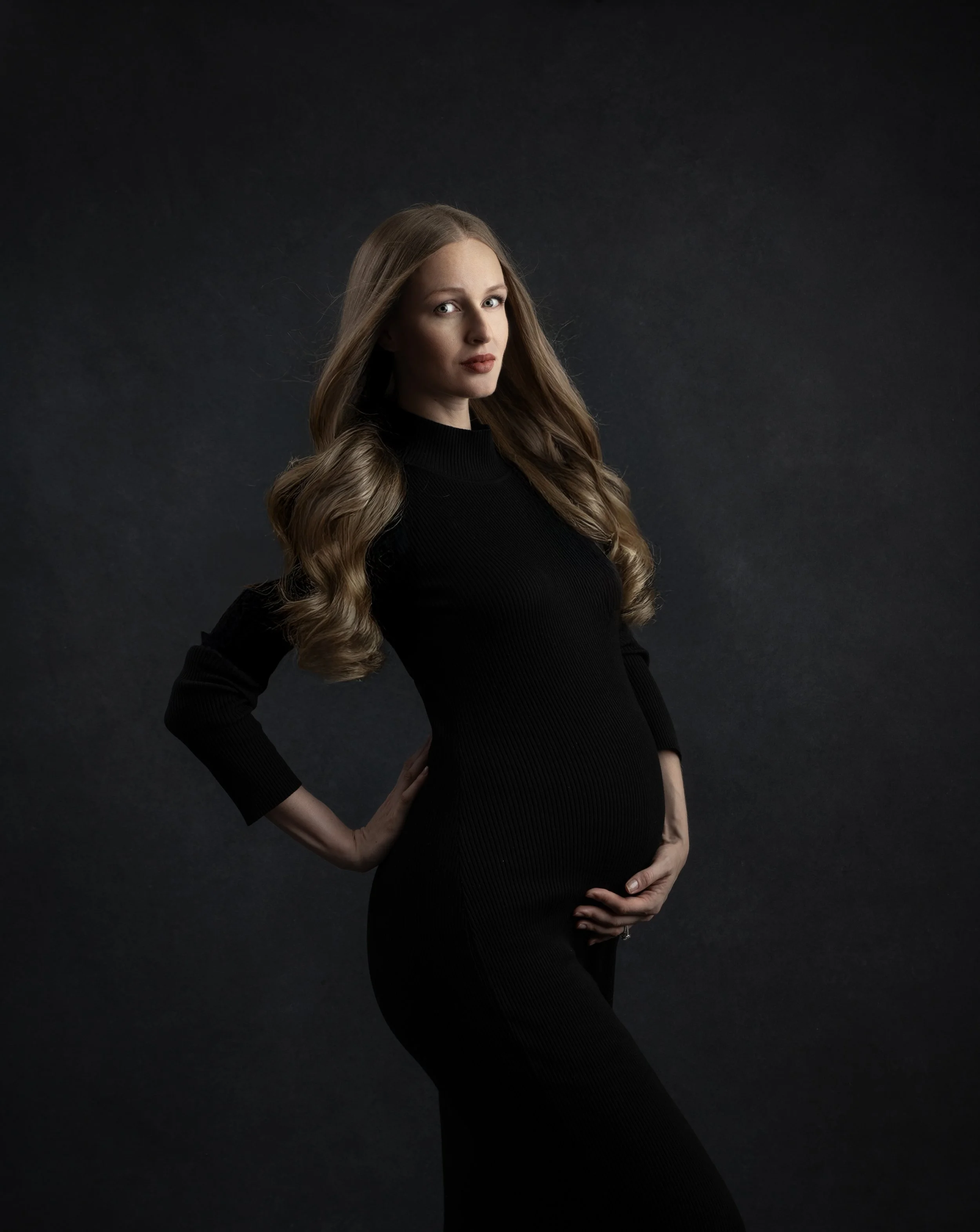 A pregnant woman with long wavy hair wearing a black dress, posing against a dark background.