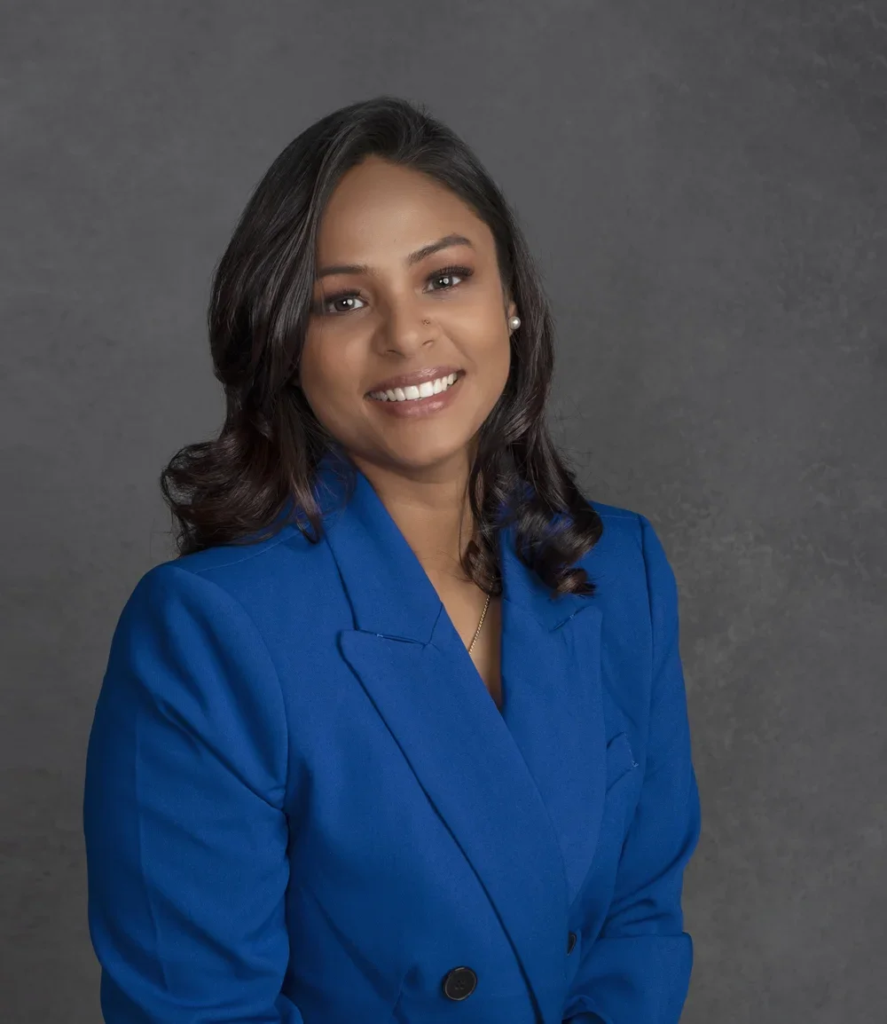 Photoshoot by Ally, A woman with dark hair, wearing a blue blazer, smiling against a gray background.