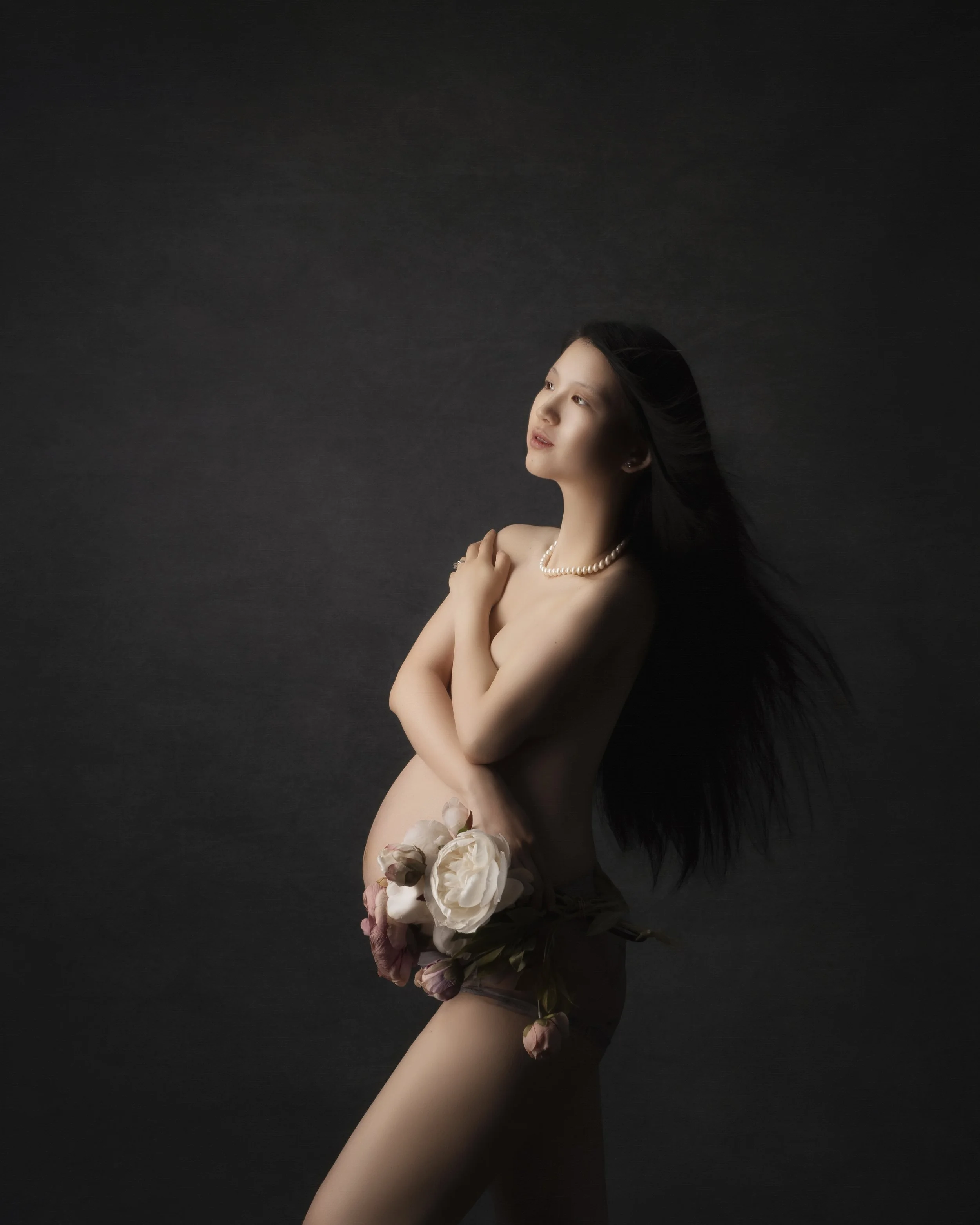 Woman with long dark hair wearing a pearl necklace, holding a bouquet of flowers, standing against a dark background.