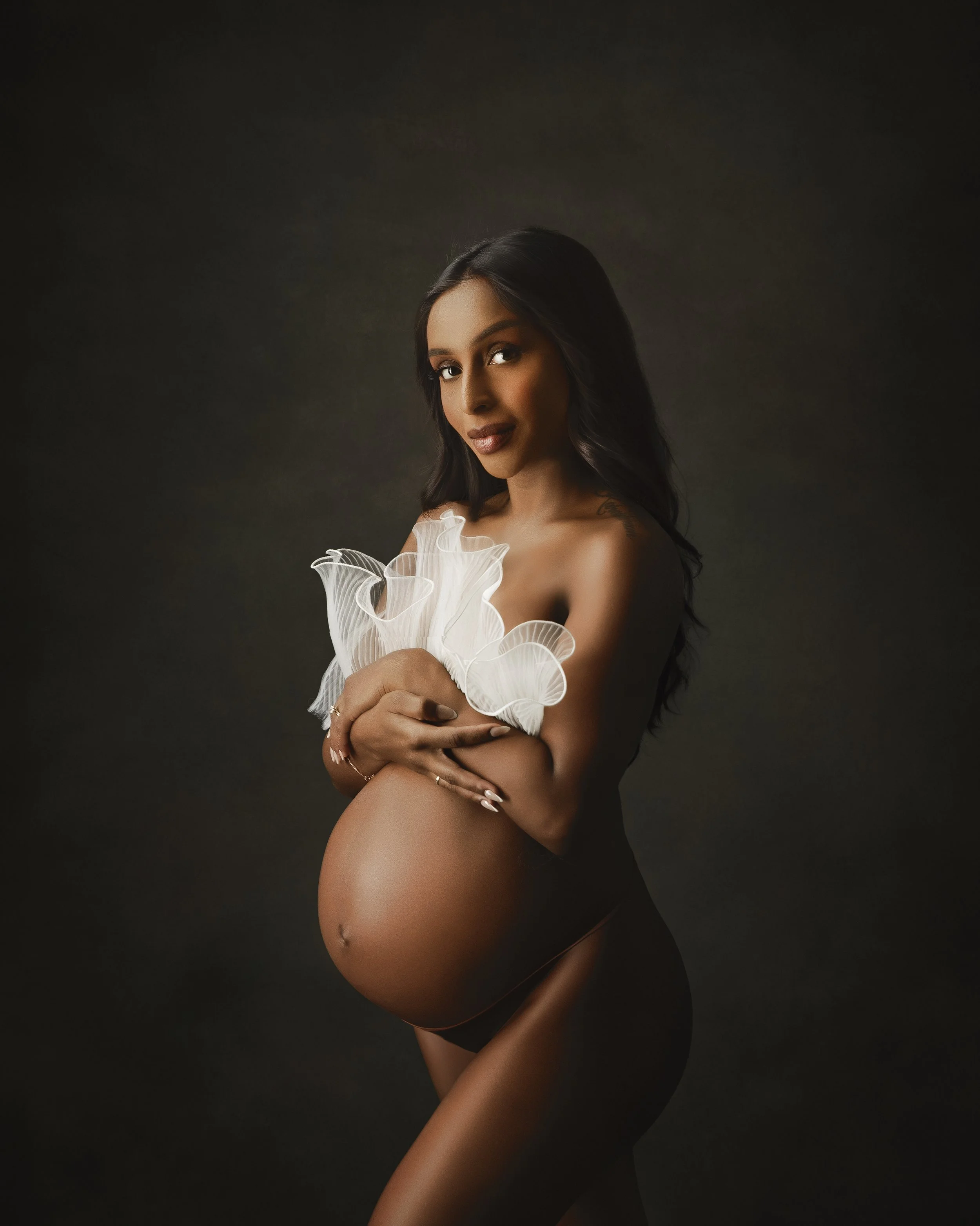 A pregnant woman with long dark hair holding a white decorative flower close to her chest against a dark background.