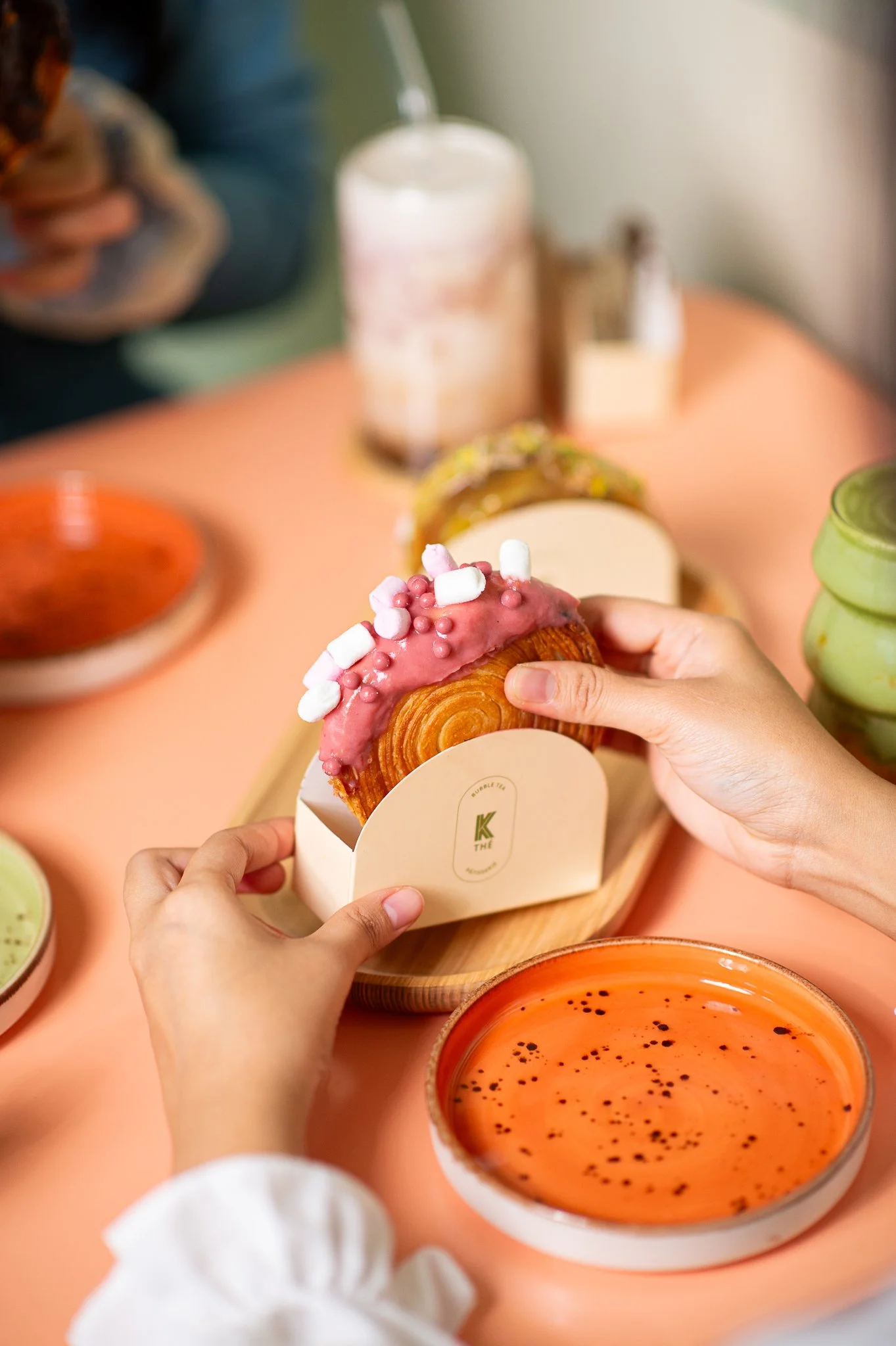 Mains tenant un pain sucré garni de glaçage rose et décorations de mini marshmallows, avec plusieurs autres desserts et boissons colorés sur la table.