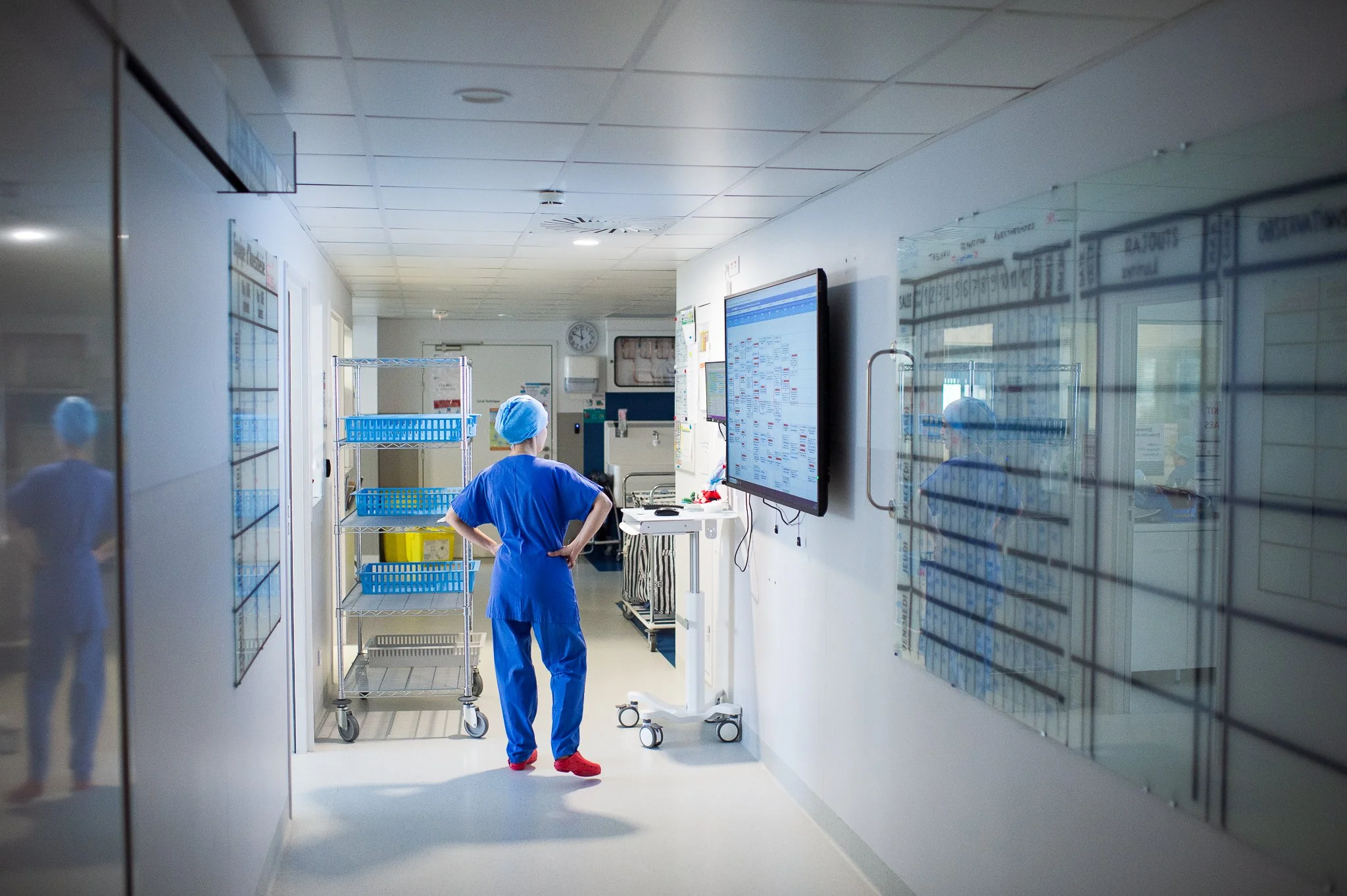Un professionnel de santé en tenue médicale bleue avec un bonnet, observant un écran dans un couloir d'hôpital.