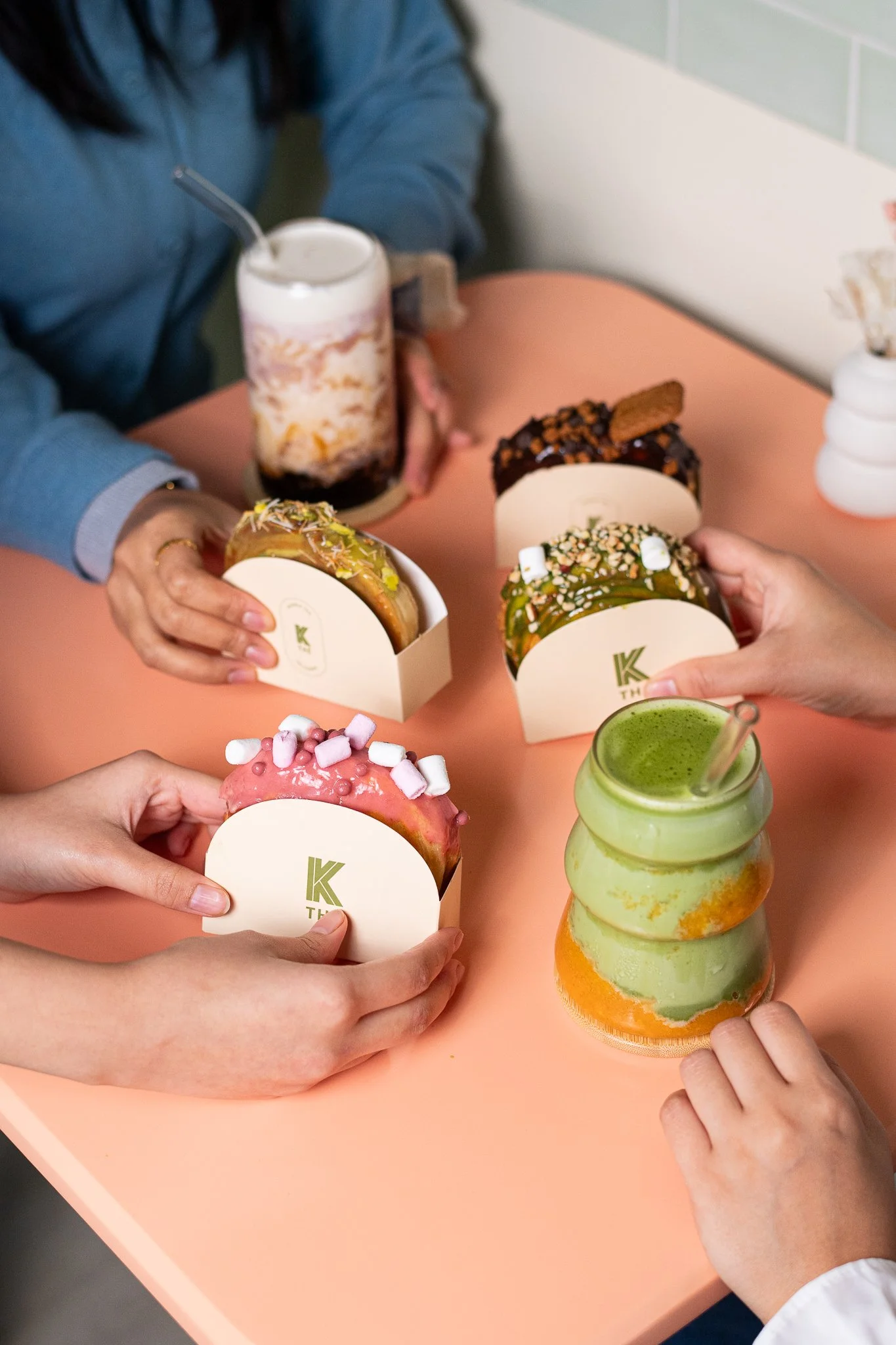 Personnes tenant des donuts décorés avec différentes garnitures et un verre de smoothie vert-orange sur une table rose.